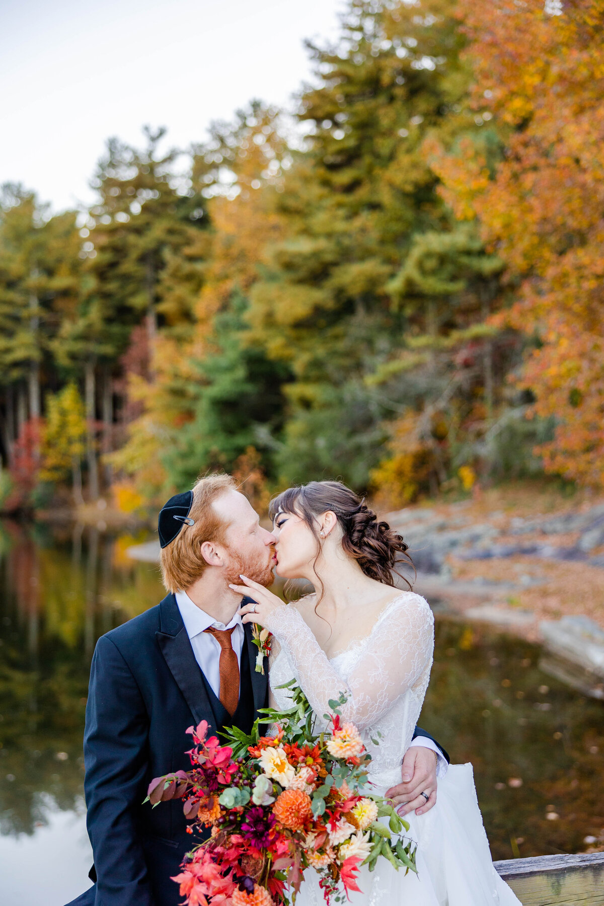 bride and groom kissing by the water with fall colors in the background