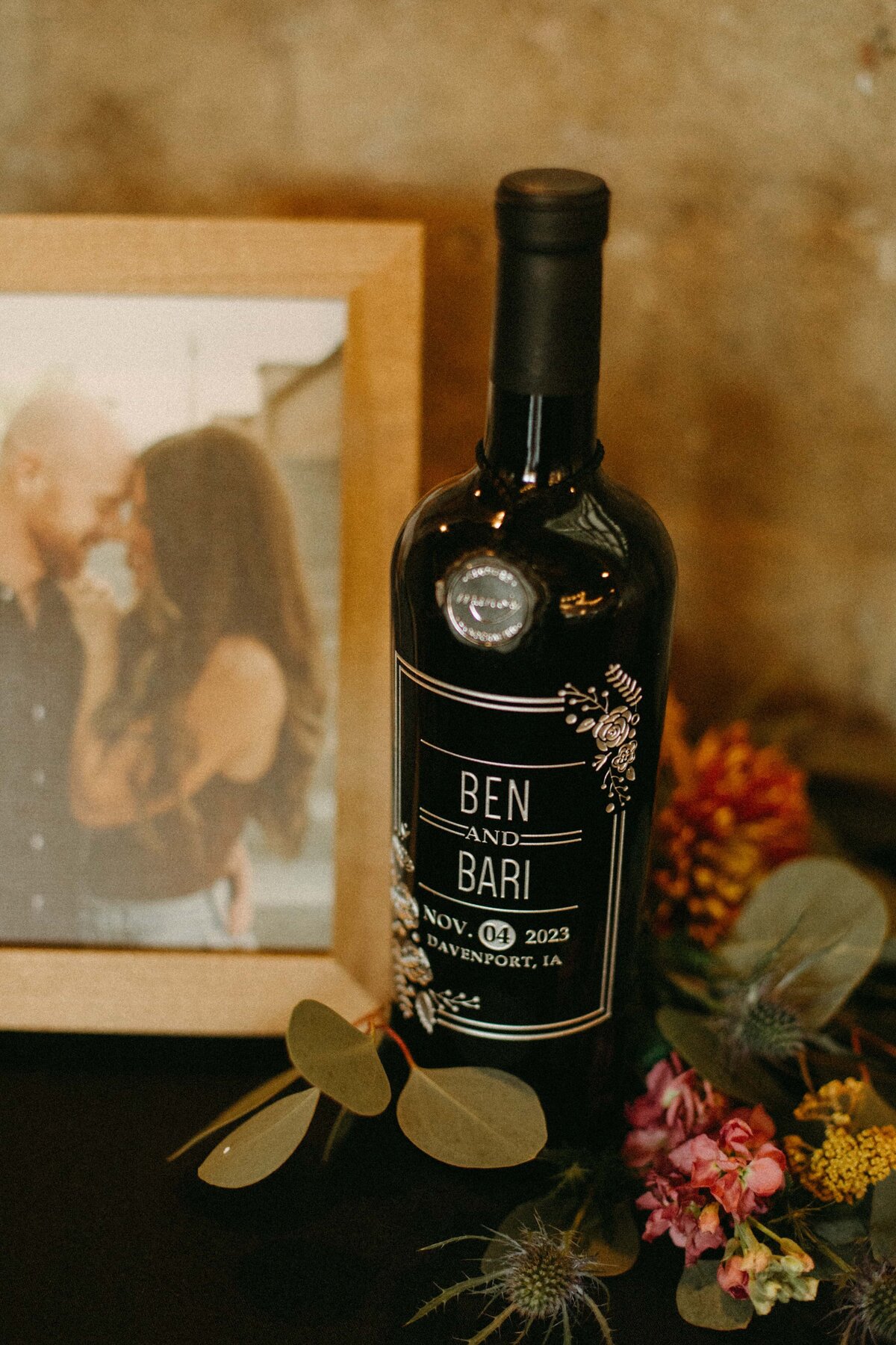 A custom-labeled wine bottle with "Ben and Bari Nov. 04, 2023" from Park Farm Winery, next to a framed photo of a couple kissing and a bouquet of
