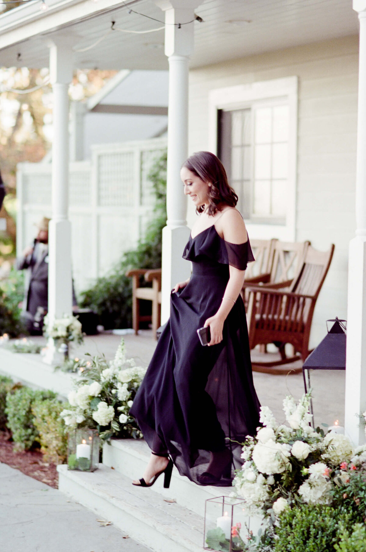 Bridesmaid gets out of a house in black off shoulder dress. Rocking wooden chairs in the patio and white flowers on the stairs.