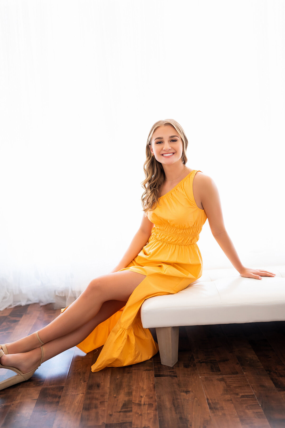 A young lady from Pewaukee High School perches on a white bench in our studio wearing a tangerine gown for her senior photos.