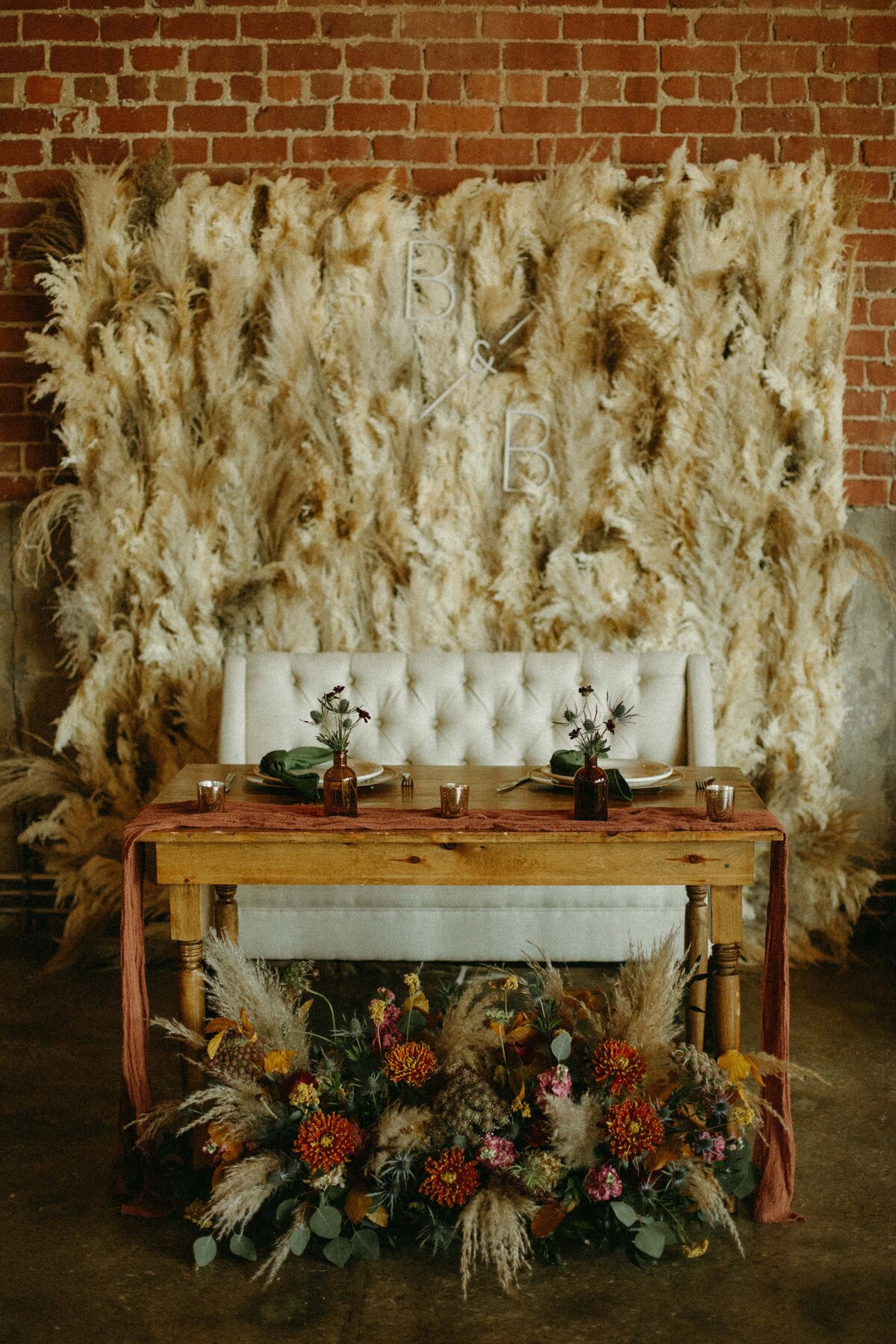 A rustic wedding reception setup at an Iowa farm featuring a wooden table with floral arrangements in front, a tufted bench, and a backdrop of feather boas with monogram initials.
