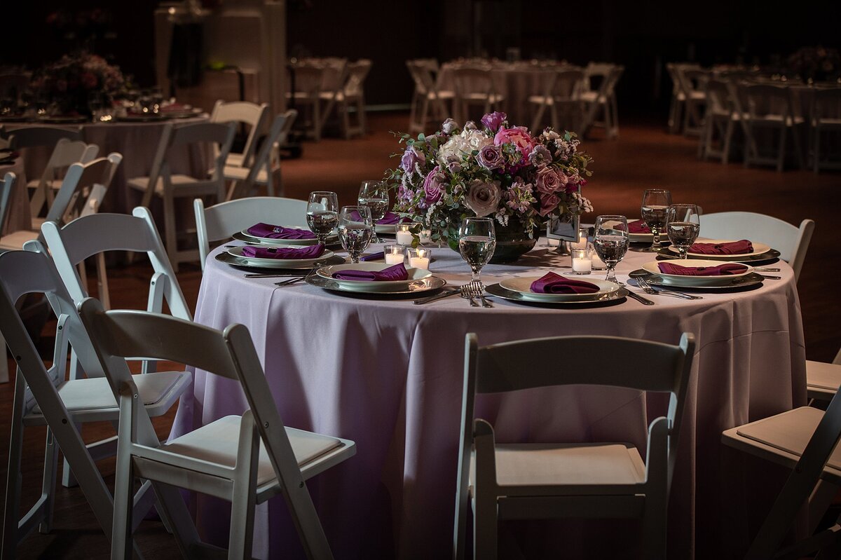 The round reception table with a light purple linen surrounded by white garden chairs  is topped with place settings of silver chargers with white plates and dark purple napkins placed at the center of each one. The floral centerpiece filled with purple roses, lilac roses, pink roses and white hydrangea is in a large silver bowl at Noah Liff Opera Center.
