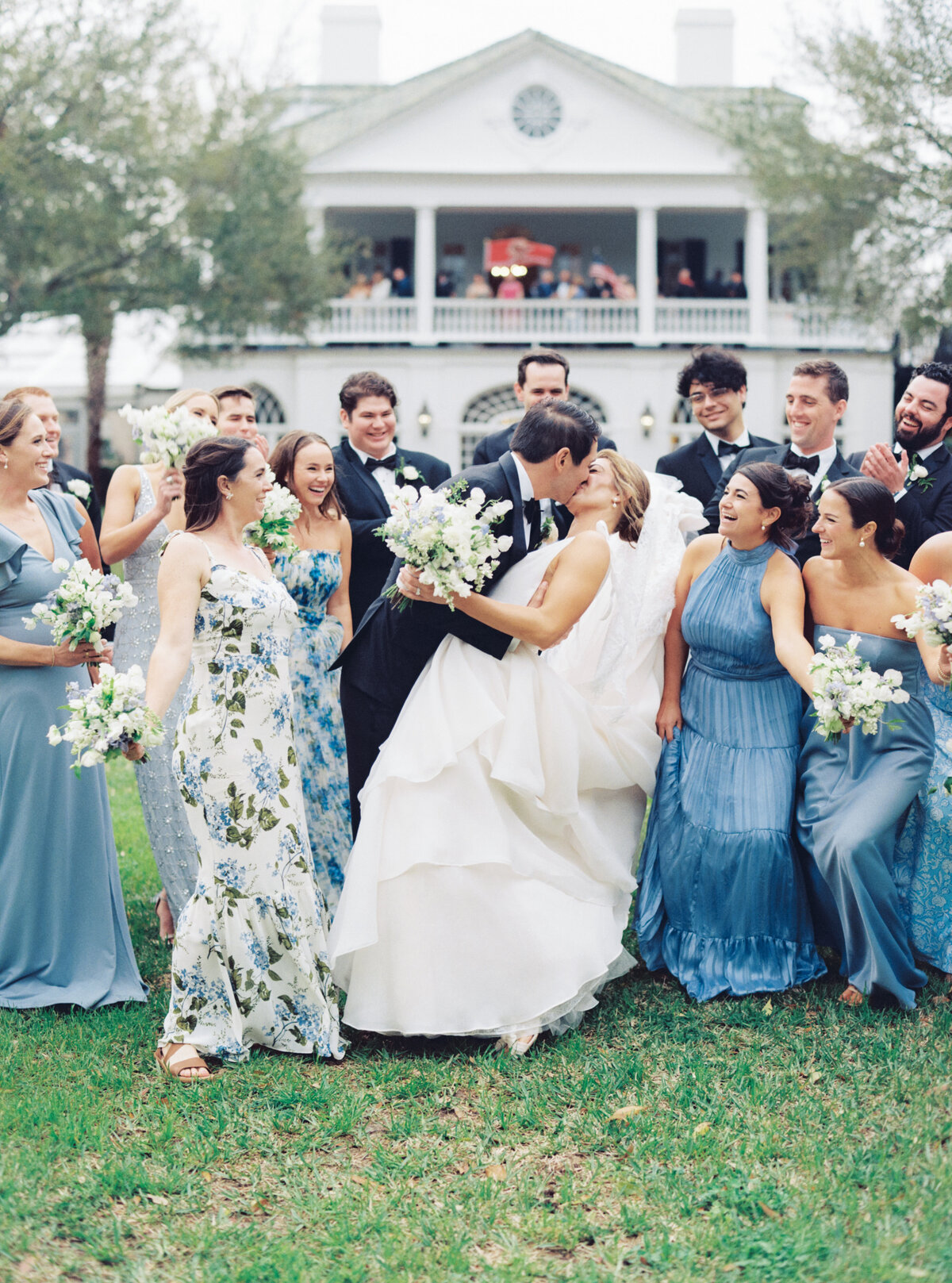 Full bridal party group photo dip kiss in front of Lowndes Grove. Bridesmaids in mix-matched blue dresses and groomsmen in classic tuxedos.
