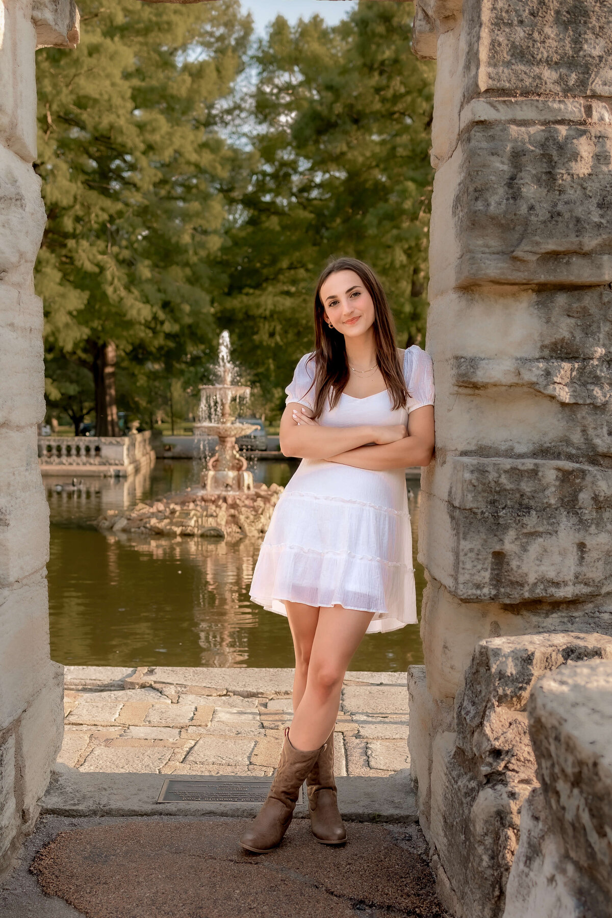 A high school senior is posing against a stone wall with a water fountain in the distance beind her.
