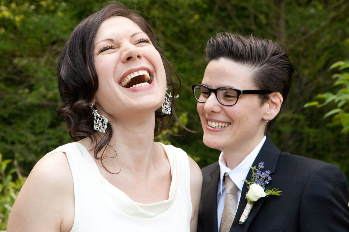 Bride in white dress laughs with her wife in wearing a dark suit lesbian New York wedding photographer Milestone Images