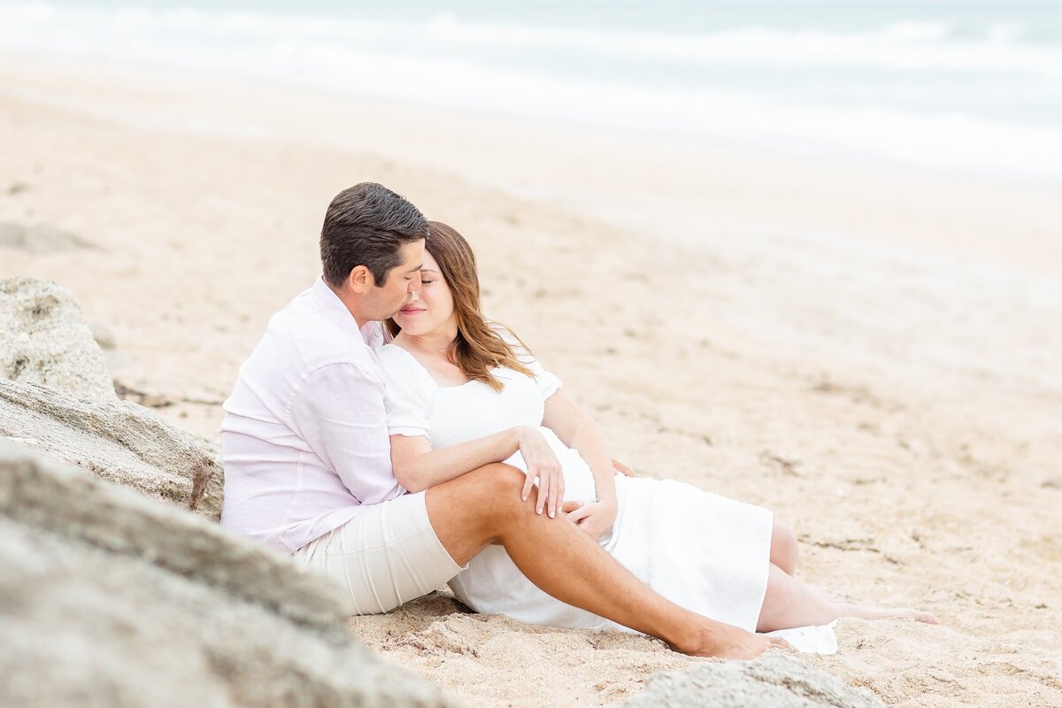 Pregnant wife snuggles in with husband sitting in the sand