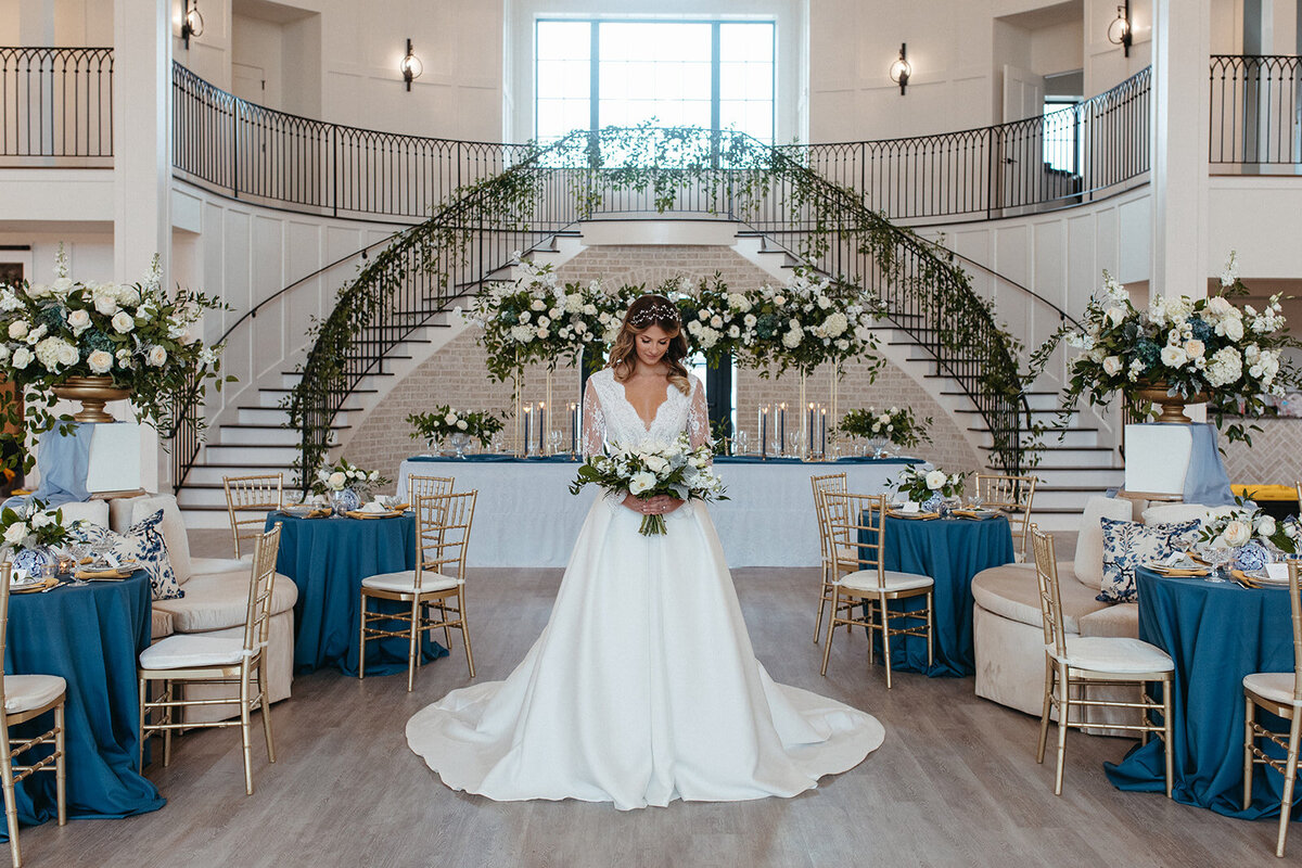 A bride in a white wedding gown looks down at her white bouquet in front of a two-sided staircase wrapped in garland.