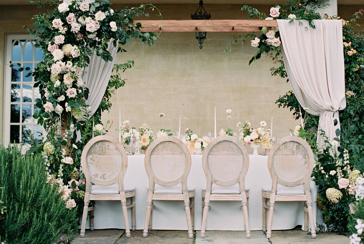 outdoor wedding table setting with blush pink flower table centrepieces and flower arch above the table