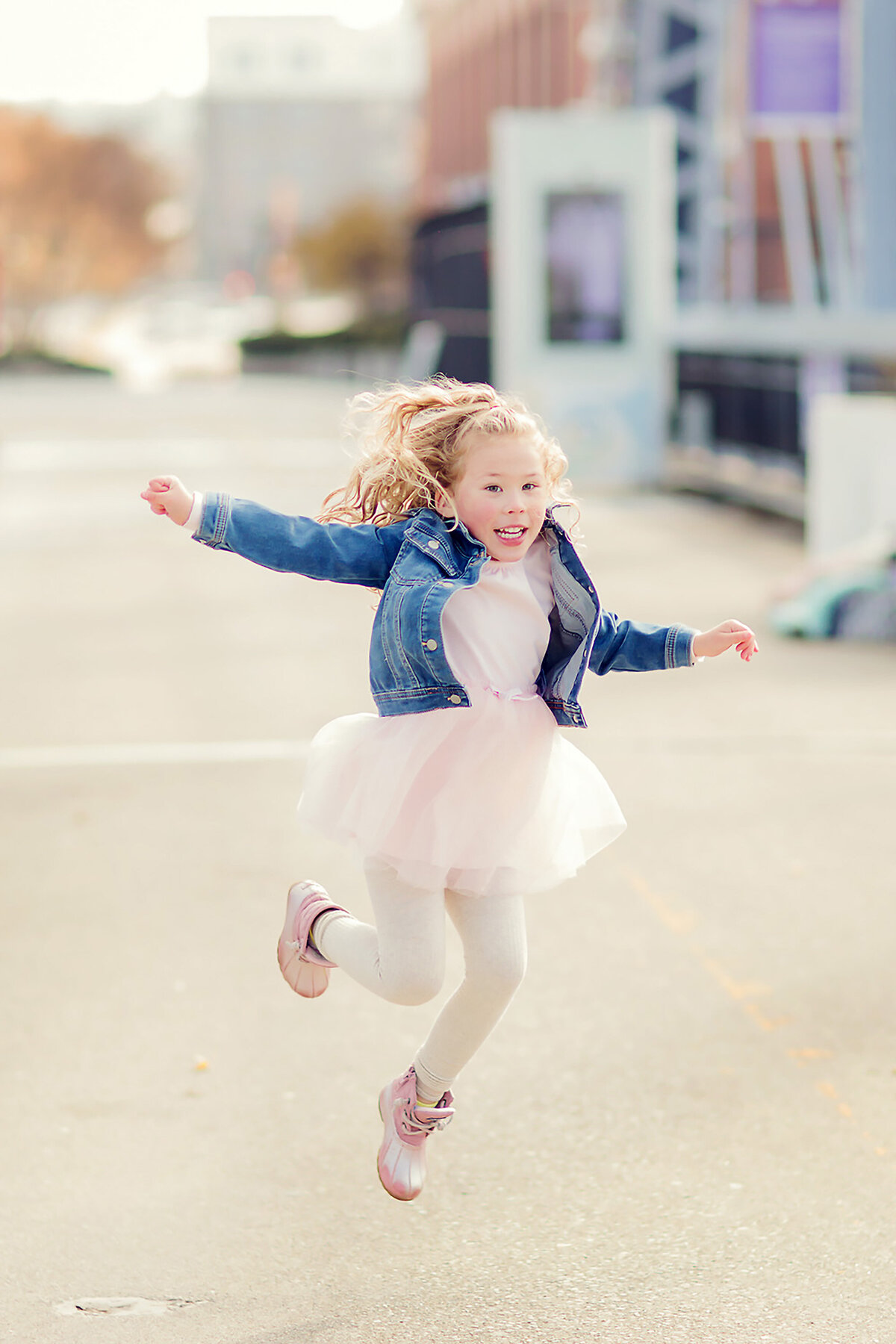 Young girl with curly blonde hair jumps gleefully in downtown Cincinnati. She's wearing a denim jacket with a tutu and pink boots.