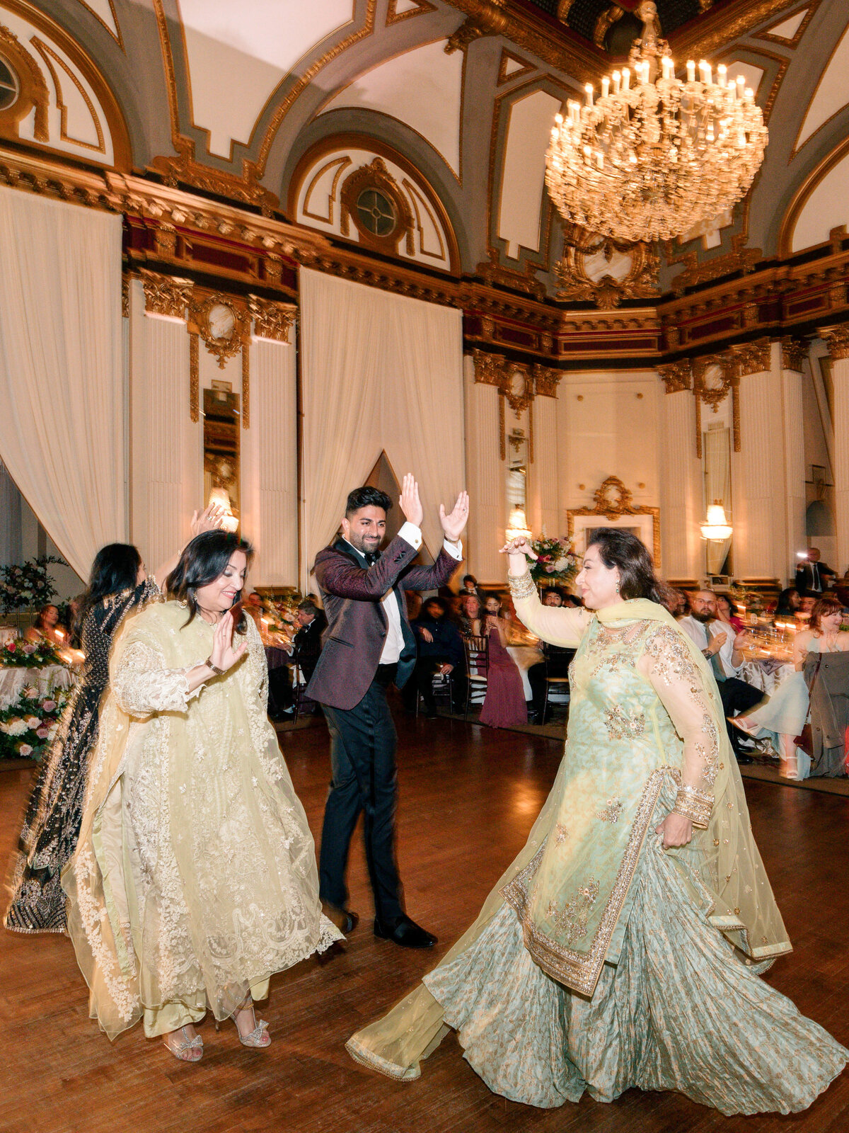 Three people dressed in elegant attire dance joyfully in a grand ballroom with chandeliers and ornate architecture. The atmosphere is lively, with seated guests and floral arrangements in the background.