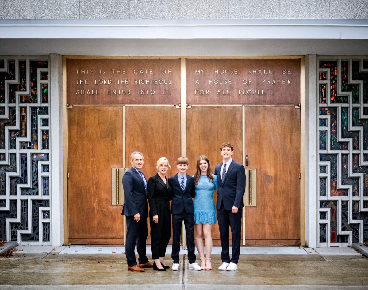 A family of five stand dressed for temple in front of the large wooden entrance doors in a garden