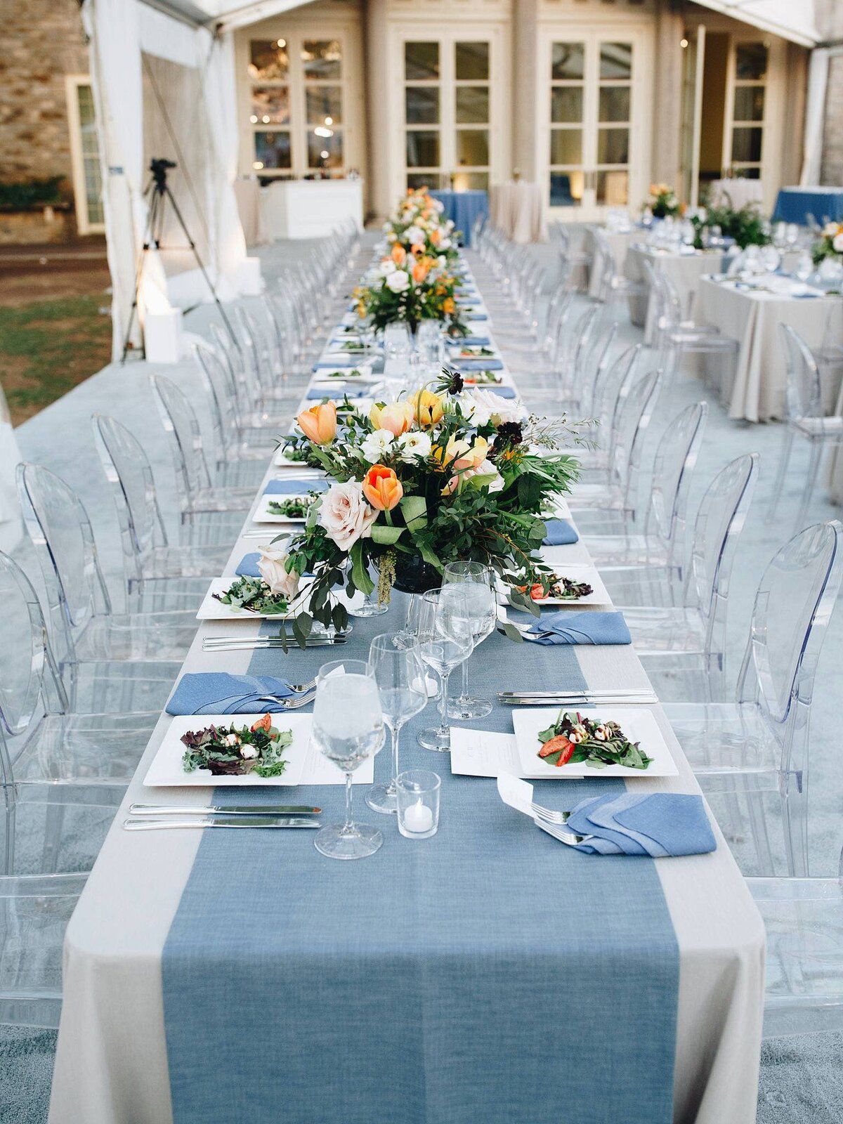 Long table lined with clear acrylic chairs. The table is covered with a long white table cloth and a wide textured table runner decorated with large centerpieces of peach, yellow and orange flowers. The tables are set with green salads on square white plates with silver flatware and a blue fancy folded napkin and long stemmed crystal glasses.