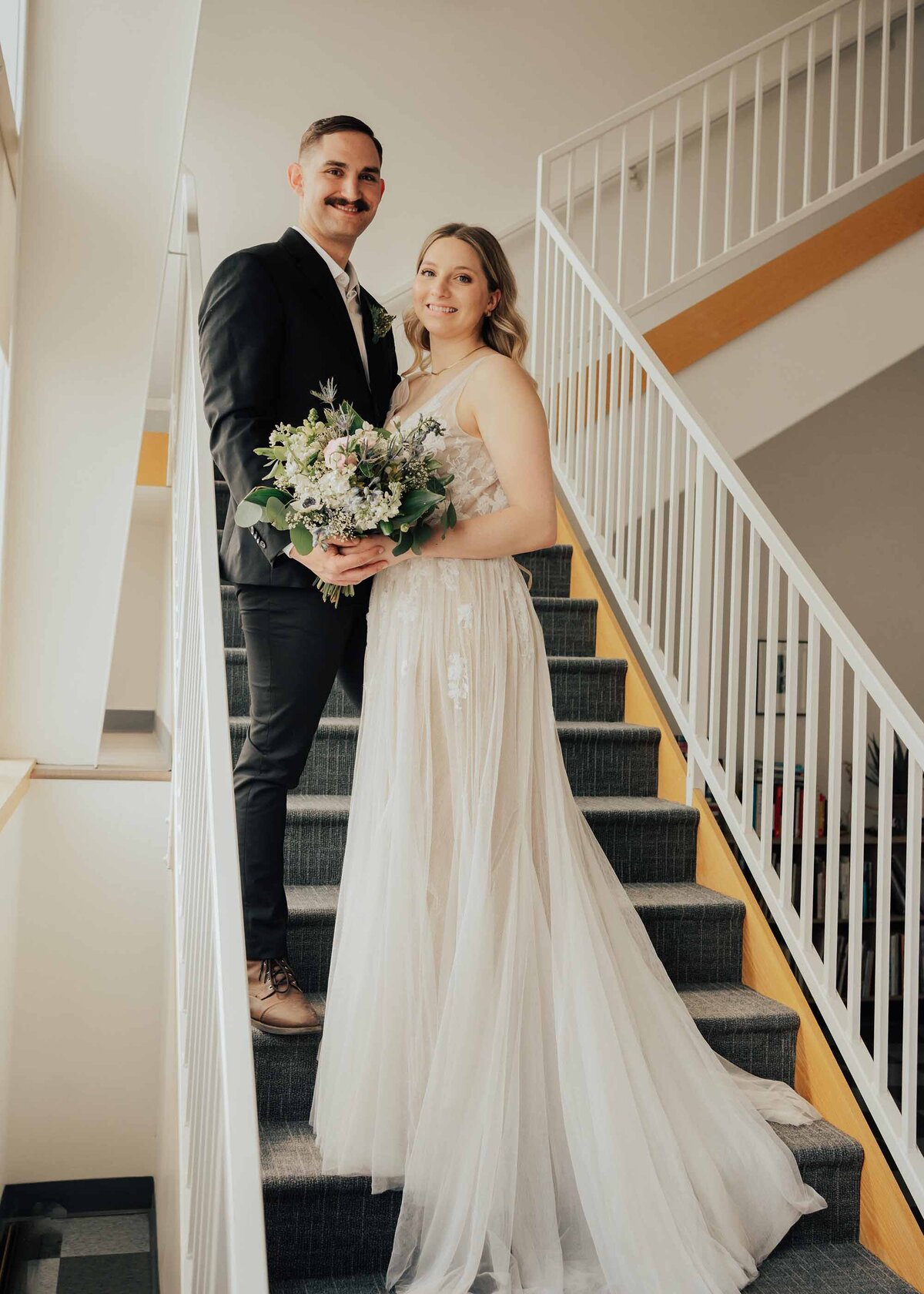 Maddie Rae Photography bride and groom standing on stairs. they are both smiling and looking at the camera. the train of her dress is flowing down the stairs