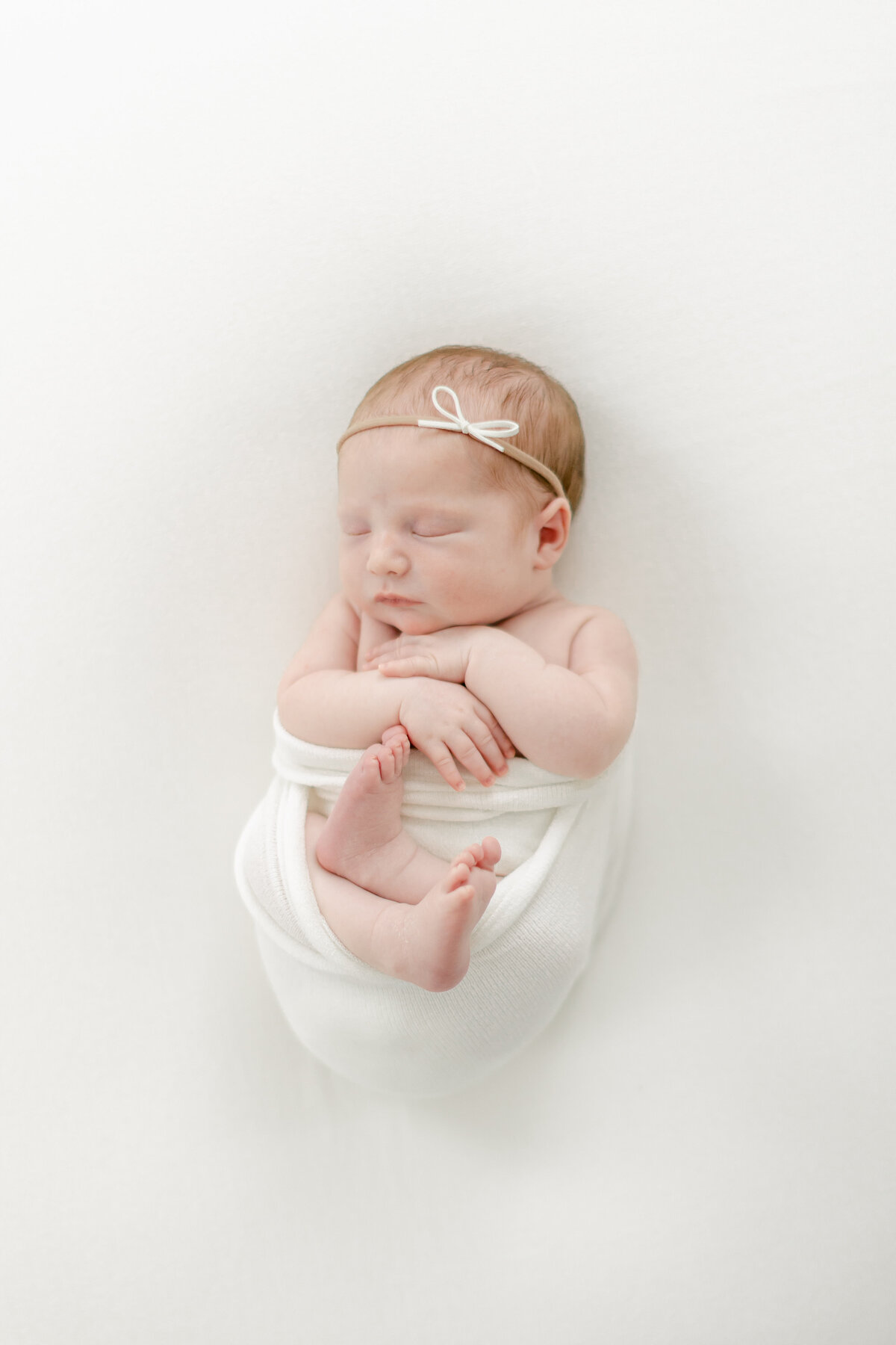 angelic newborn baby girl wrapped in white swaddle wearing a dainty headband for her newborn session photographed by Philadelphia Newborn Photographer Tara Federico