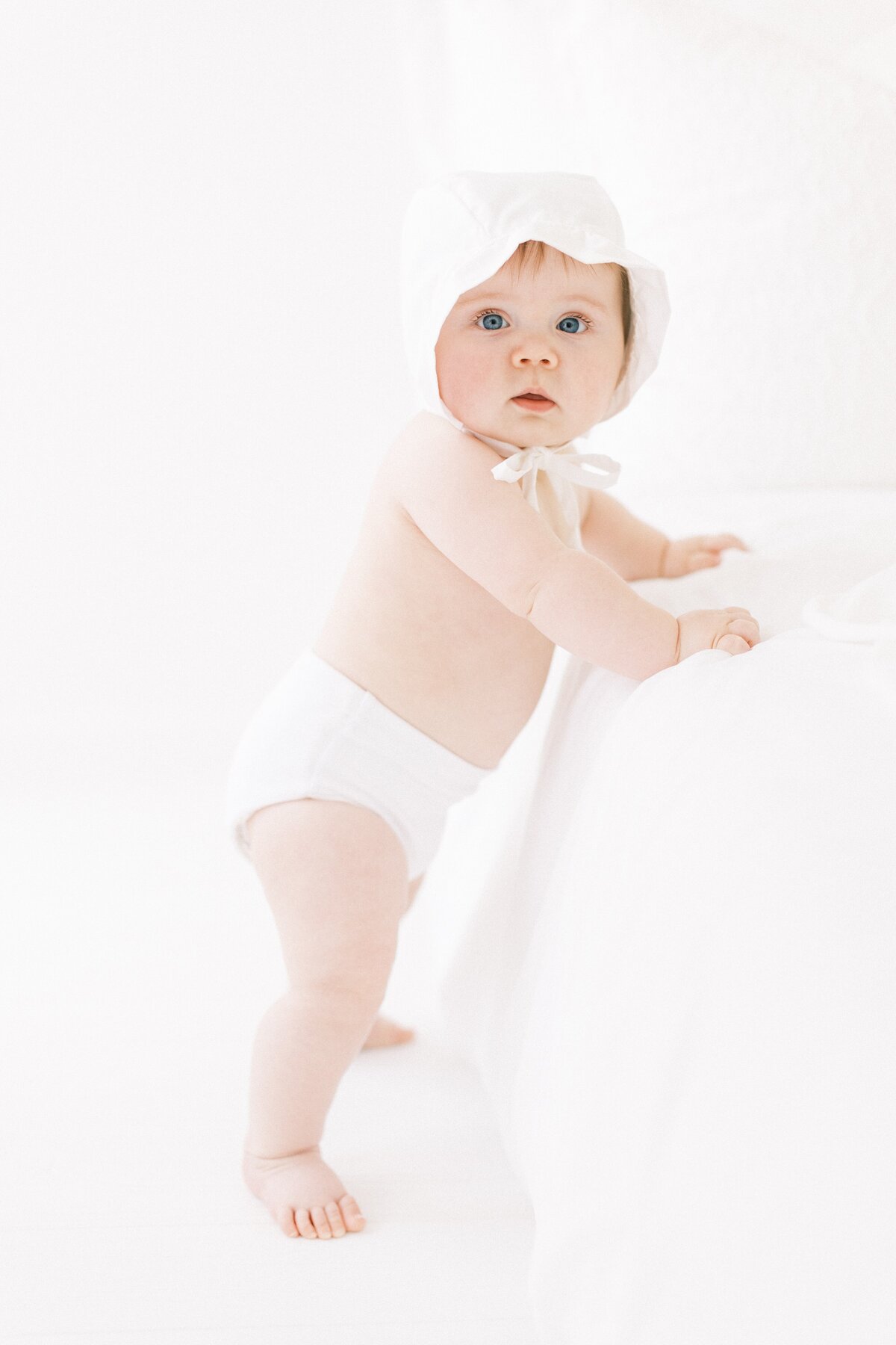 6 month old baby girl wearing a white bonnet standing and looking at camera during baby studio session