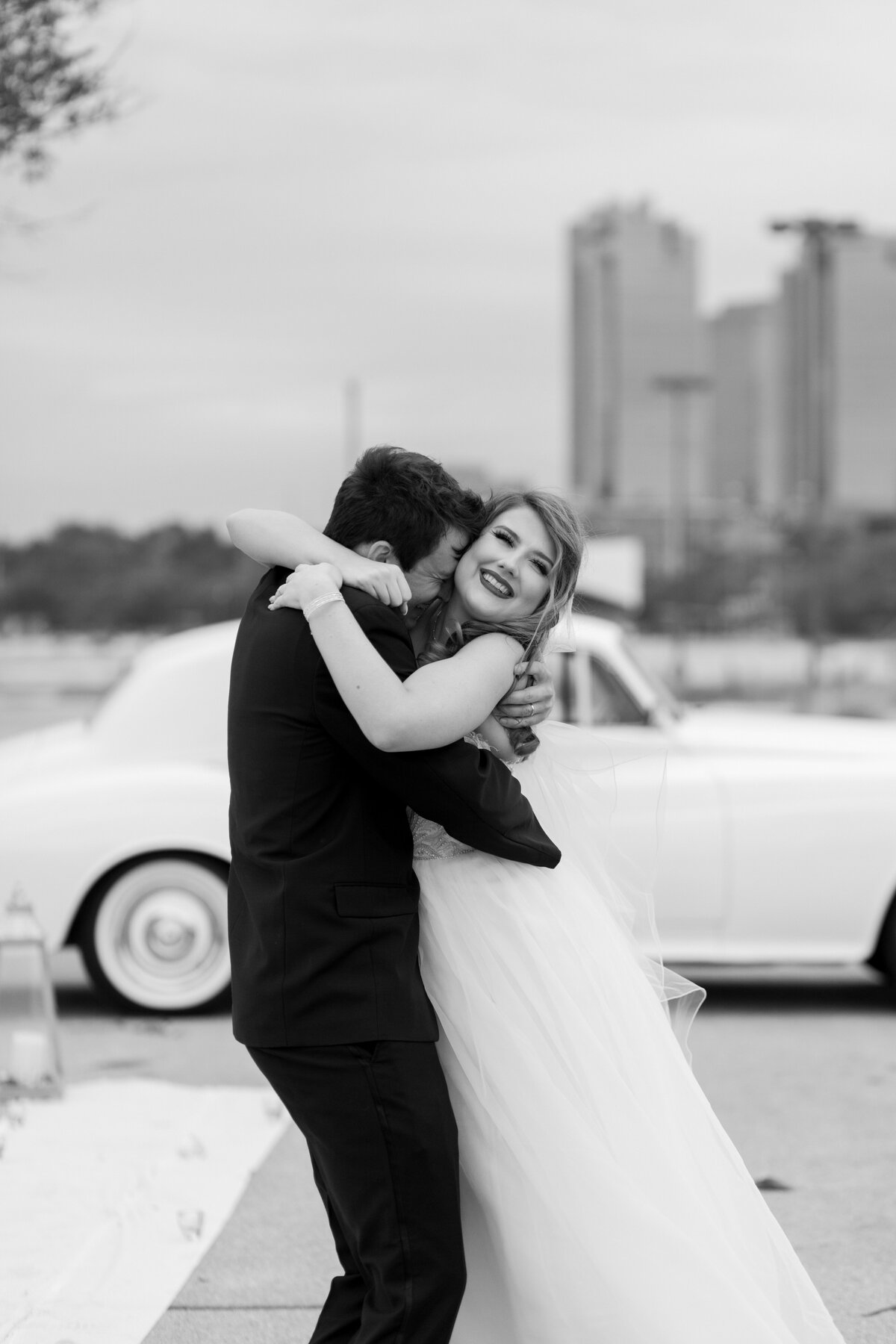 A black and white candid photo of a groom deeply embracing his bride after their elopement in Fort Worth, Texas. The groom is on the left and is wearing a black suit and has his face buried into the bride's neck. The bride is wearing a flowing, white dress, and is looking towards the camera with a large smile on her face. An antique car is behind them with the Fort Worth skyline framing the scene.
