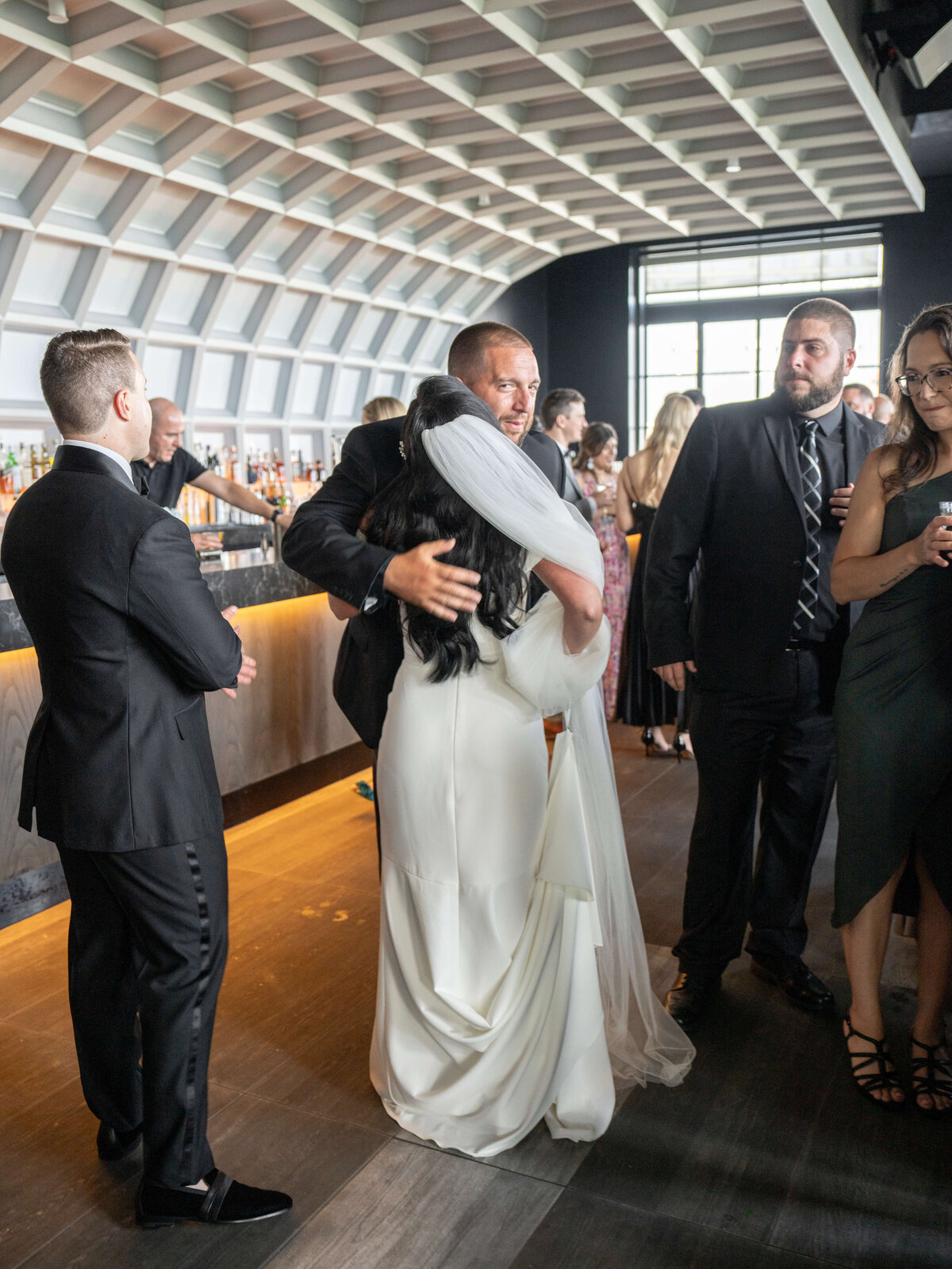 A bride and her wedding guest share a hug at a cocktail hour held in a modern venue with a curved ceiling. The woman is in a white dress, and the man is in a suit. Other guests in suits and dresses are mingling nearby.