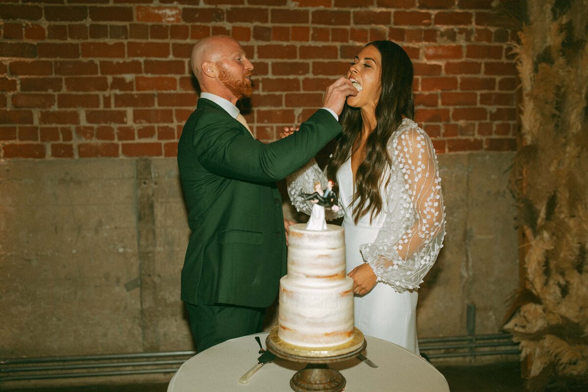 A bald man in a green suit feeds wedding cake to a smiling woman in a white lace dress at an Iowa wedding in front of a brick wall.