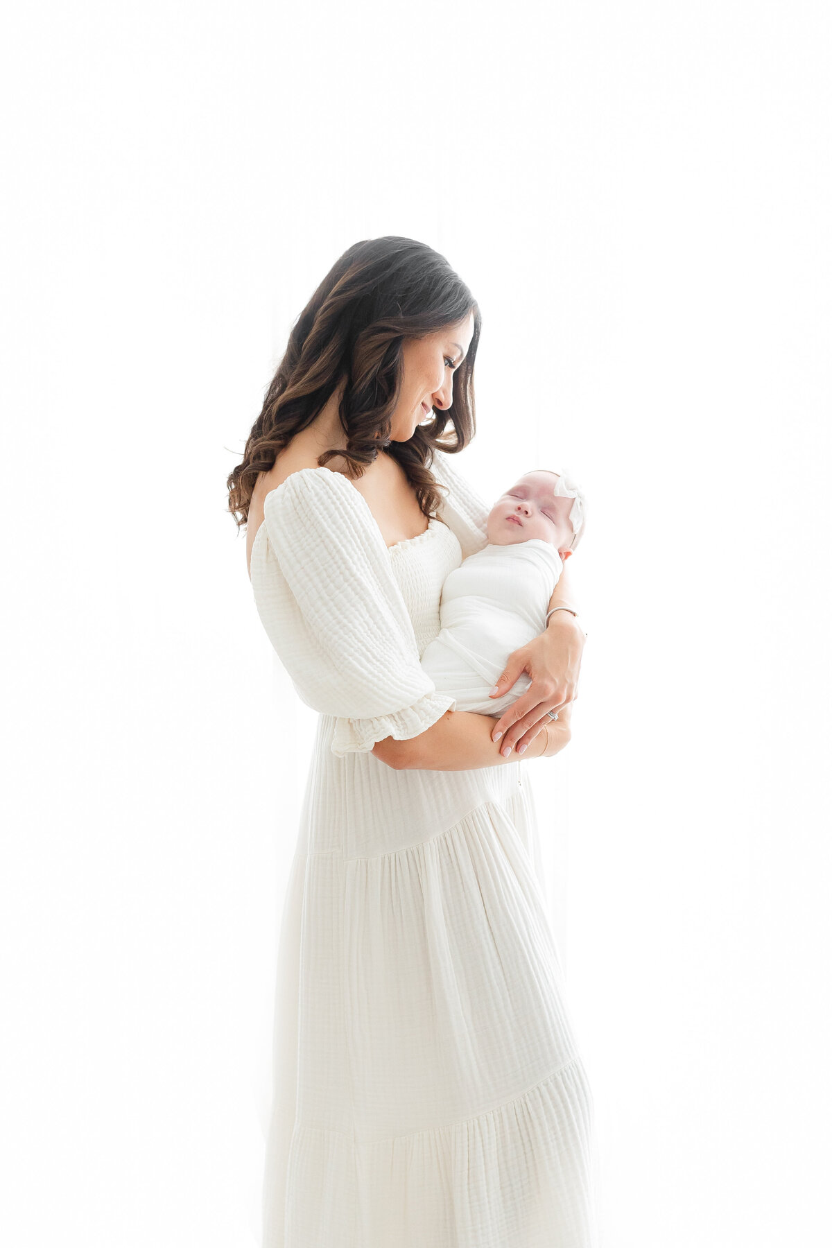A bright white DC Newborn Photography photo of a mother wearing a long white waffle knit dress in front of a window with her newborn baby girl