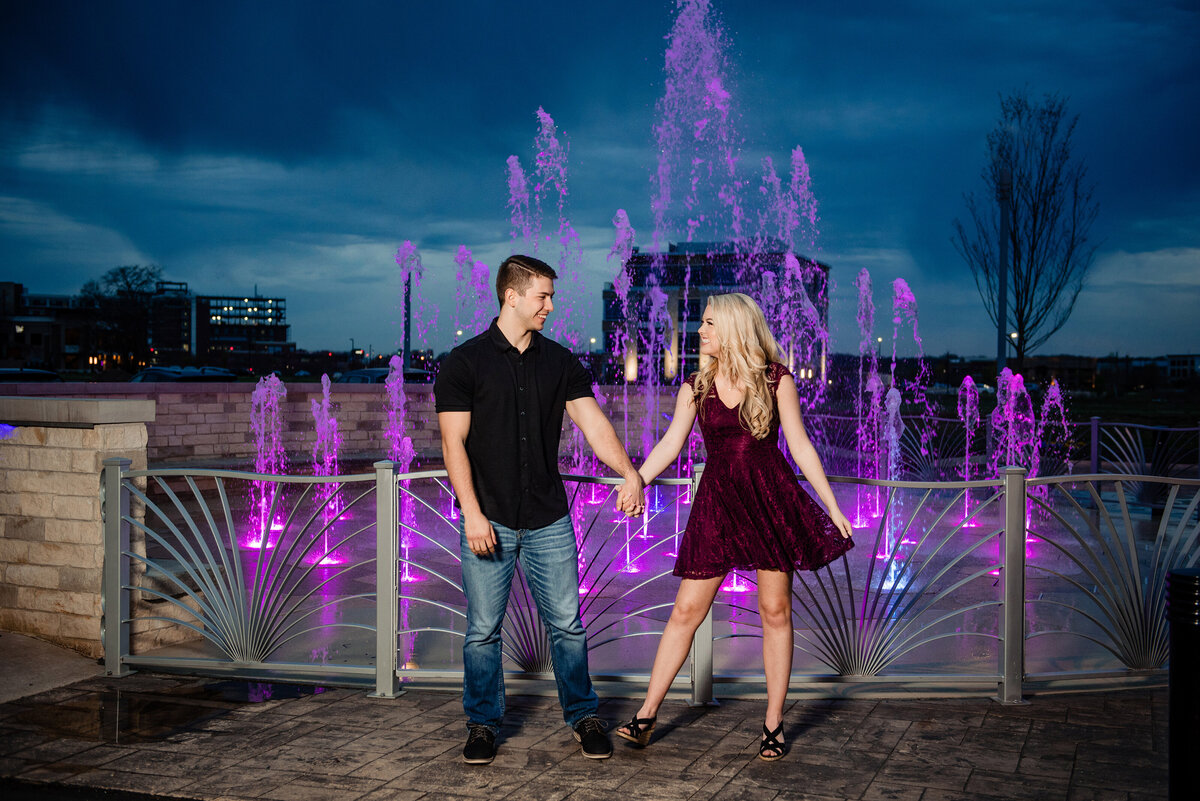 Engaged couple laughing hand in hand with purple fountain behind them