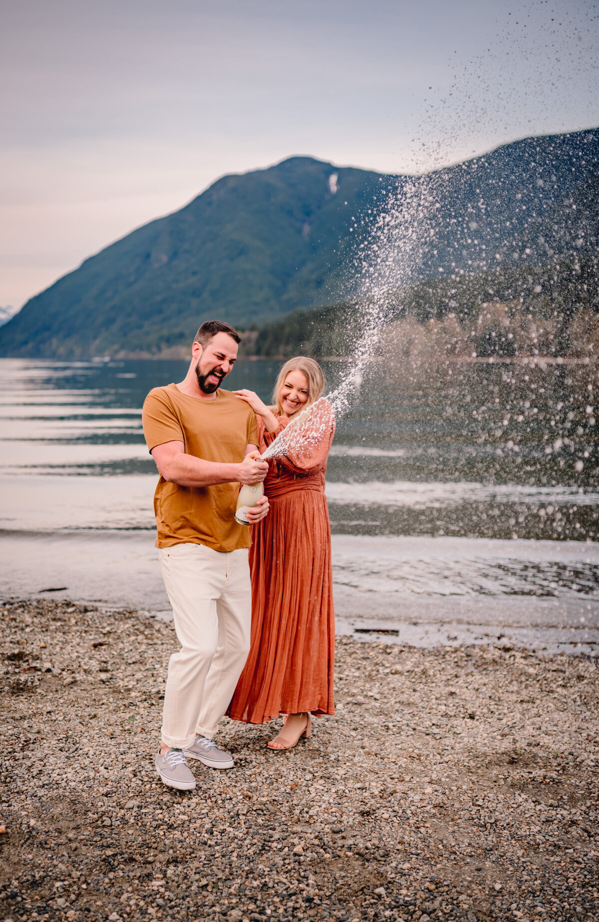 Newly engaged couple holding champagne bottle and champagne falling out to celebrate engagement. Couple dressed in white and light orange, mountain and lake in the back round. 