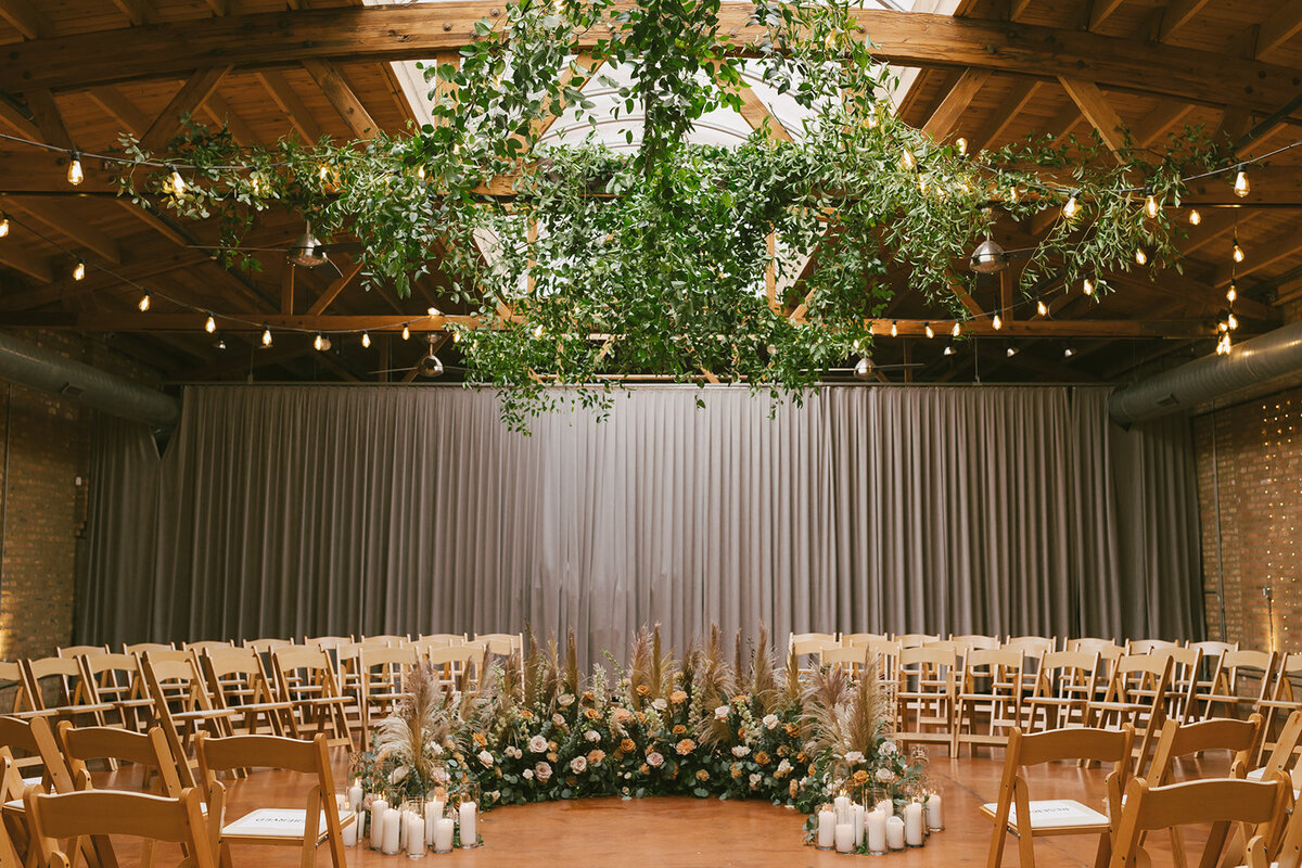 Several wooden chairs in a circle surround lush greenery, pampas grass, candles and white flowers before ceremony at Loft on Lake, Chicago.