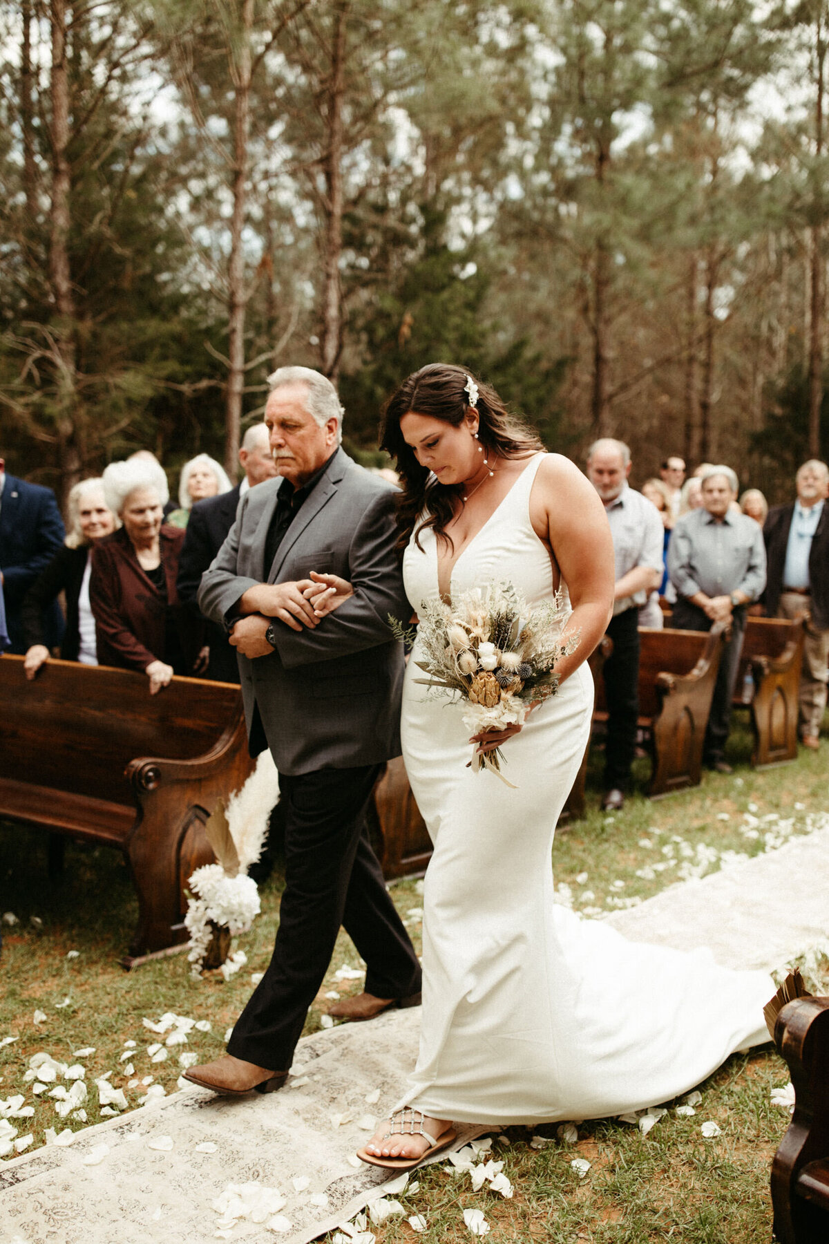Father walking his daughter down the aisle during an outdoor ceremony in the woods with vintage rugs under them and wooden pews behind them