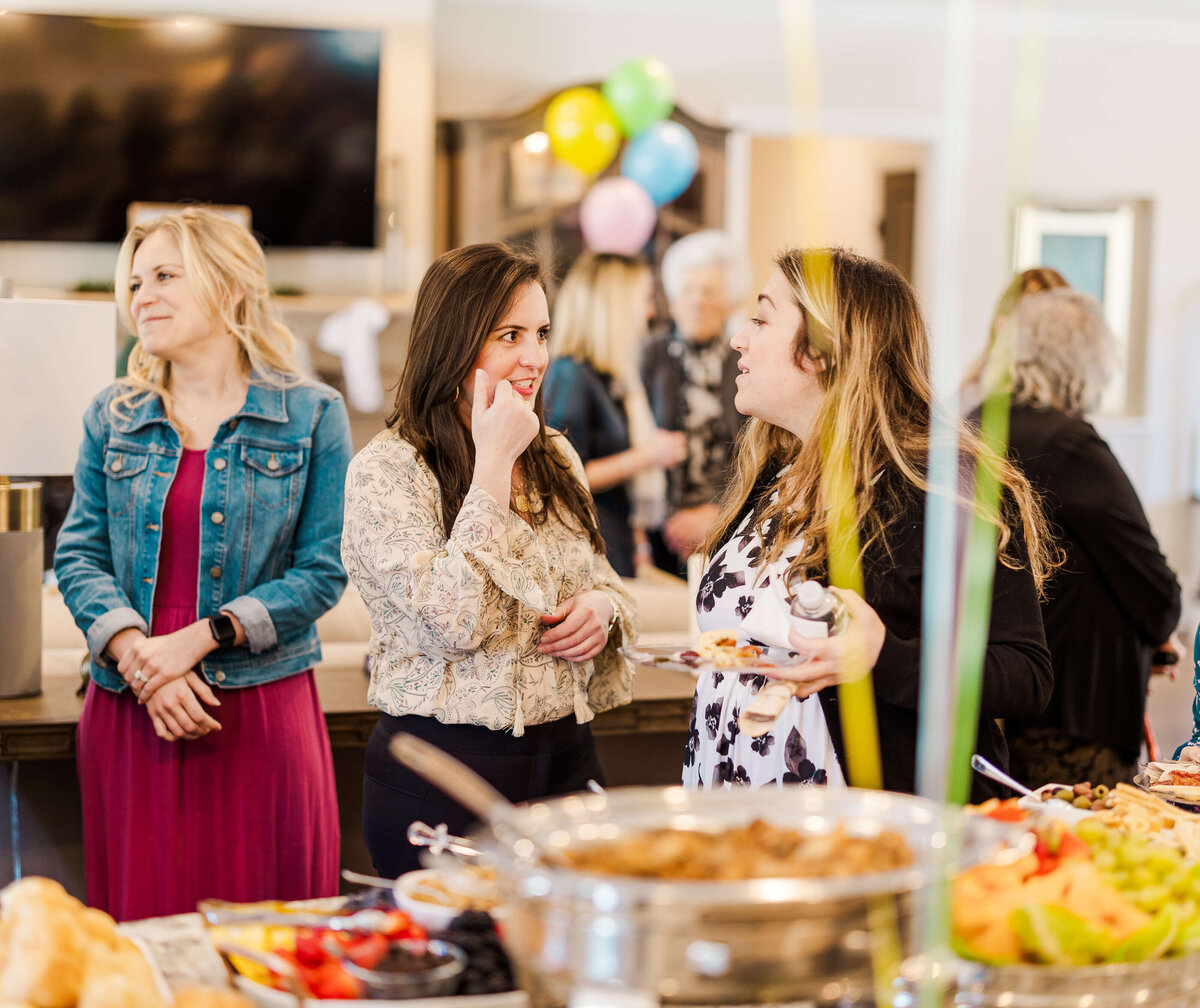 Three women are standing at a buffet table, conversing. The table is filled with various dishes, including fruit and a heated chafing dish. Balloons in the background suggest a festive gathering captured by an event photographer in Fayetteville, NC.