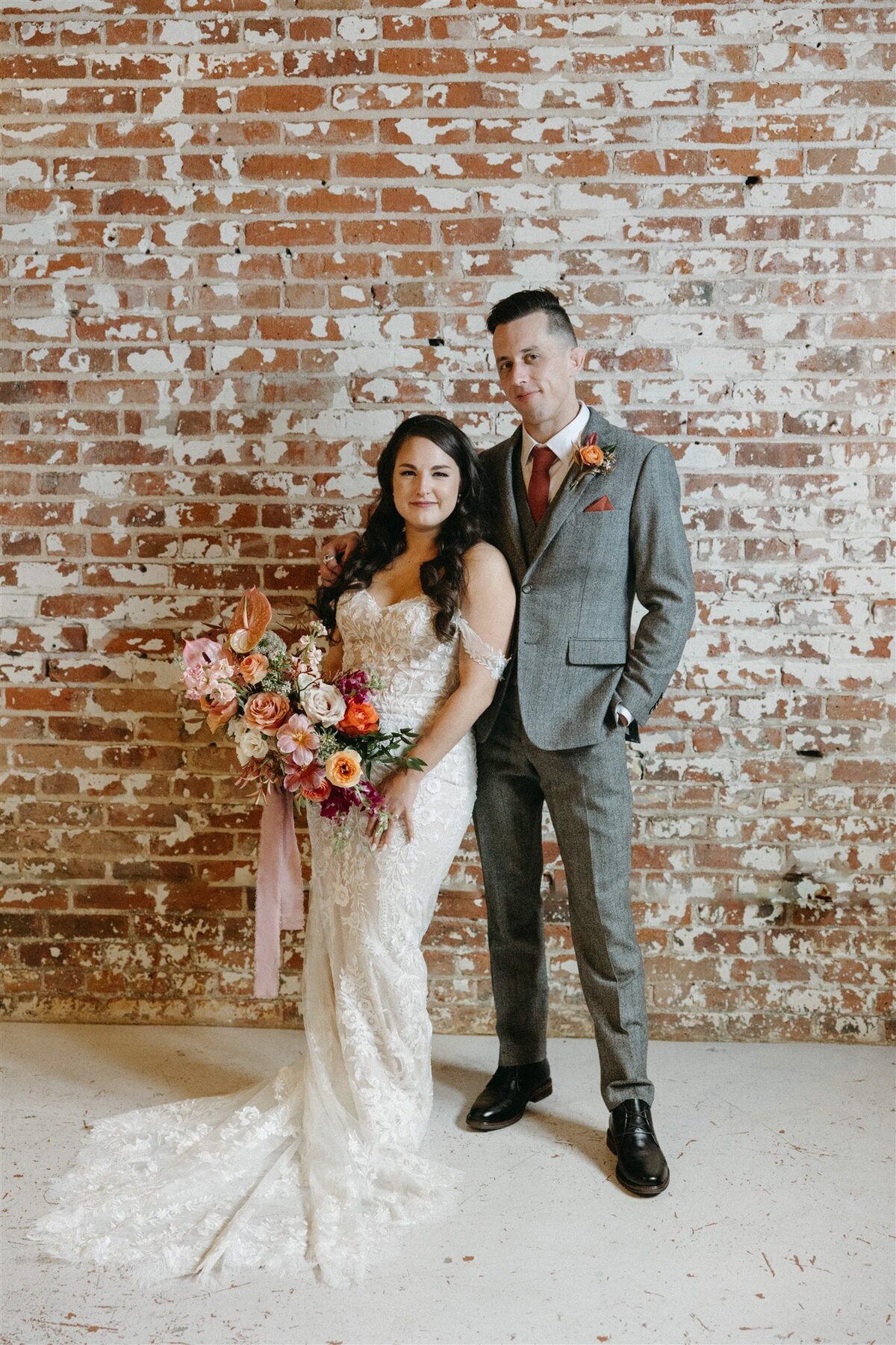 Bride wearing lace dress holding colorful bouquet and groom  wearing grey suit standing against brick wall  at the St Vrain, Longmont CO.