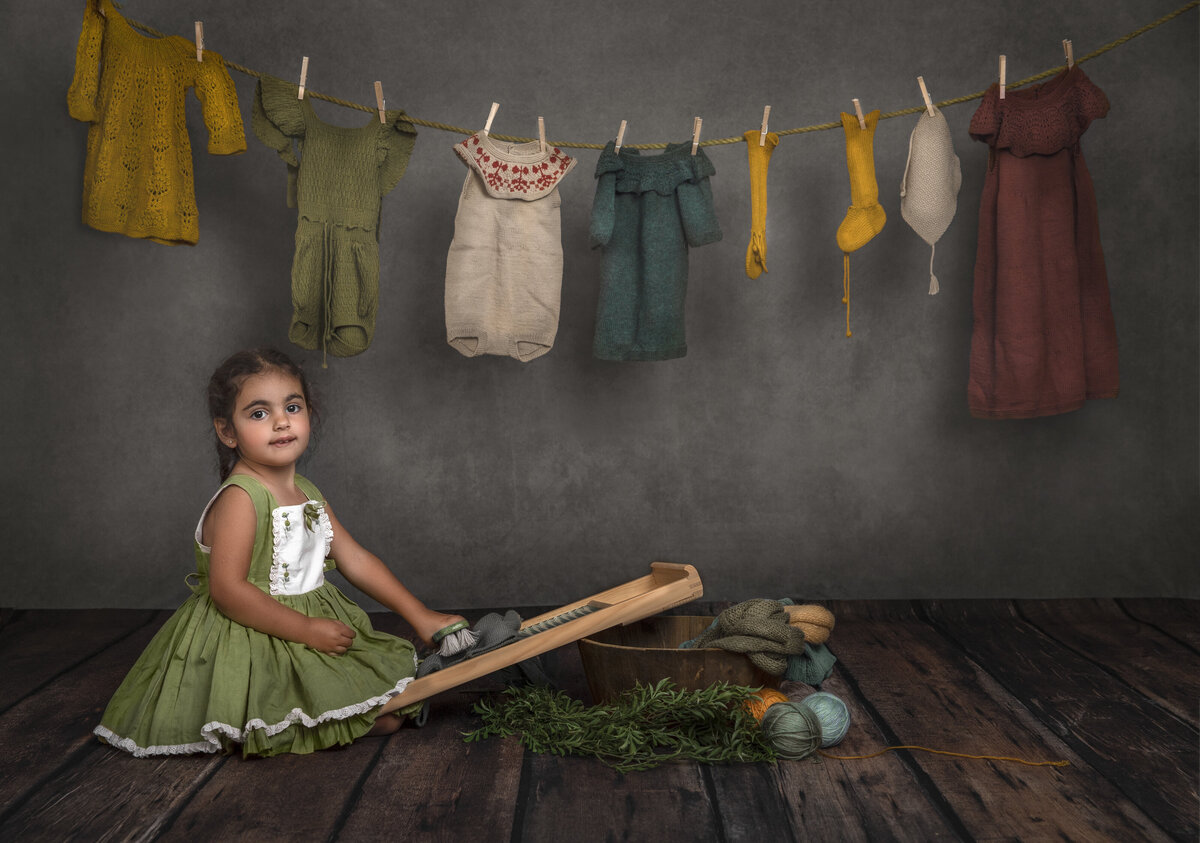 A young girl sits beneath a line of clothes' dressed in a green  dress.  she is sitting with a board resting on top of a basket of yarn.