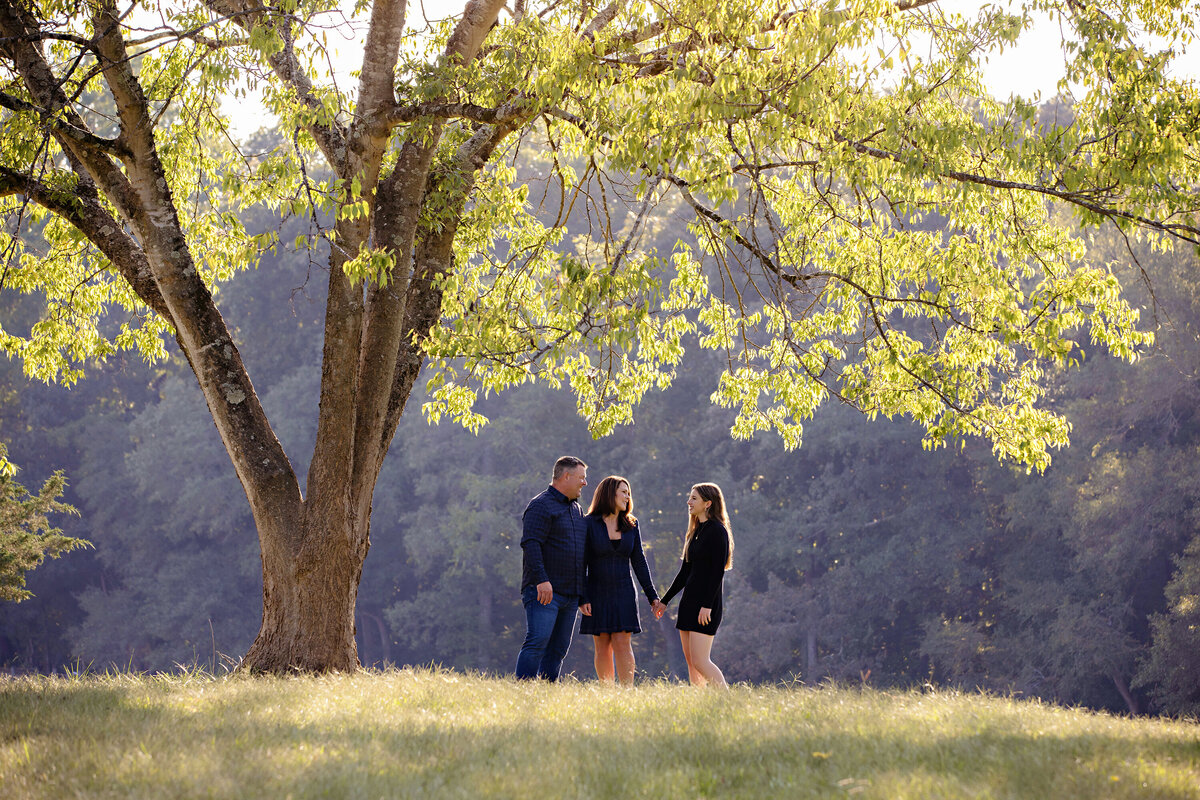 Mom, Dad and teen daughter photographed looking at each other off in the distance by Carolyn Ann Ryan. The family is standing underneath the canopy of a stunning huge tree and the leaves are all backlit in a bright green with the shadowed backdrop of darker trees behind them in the distance. 