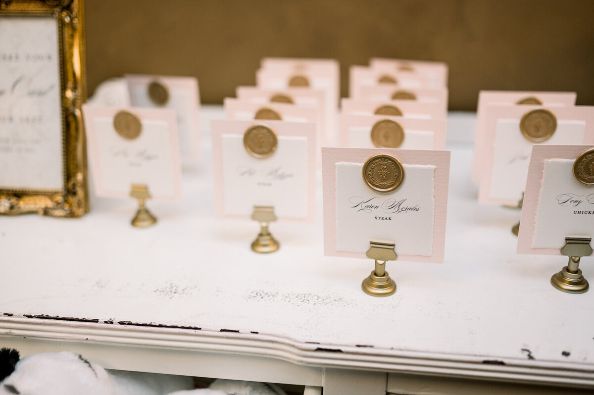 Blush escort cards made of hand-torn paper with gold wax seals displayed in gold stands on a white buffet table for a 50th wedding anniversary celebration.
