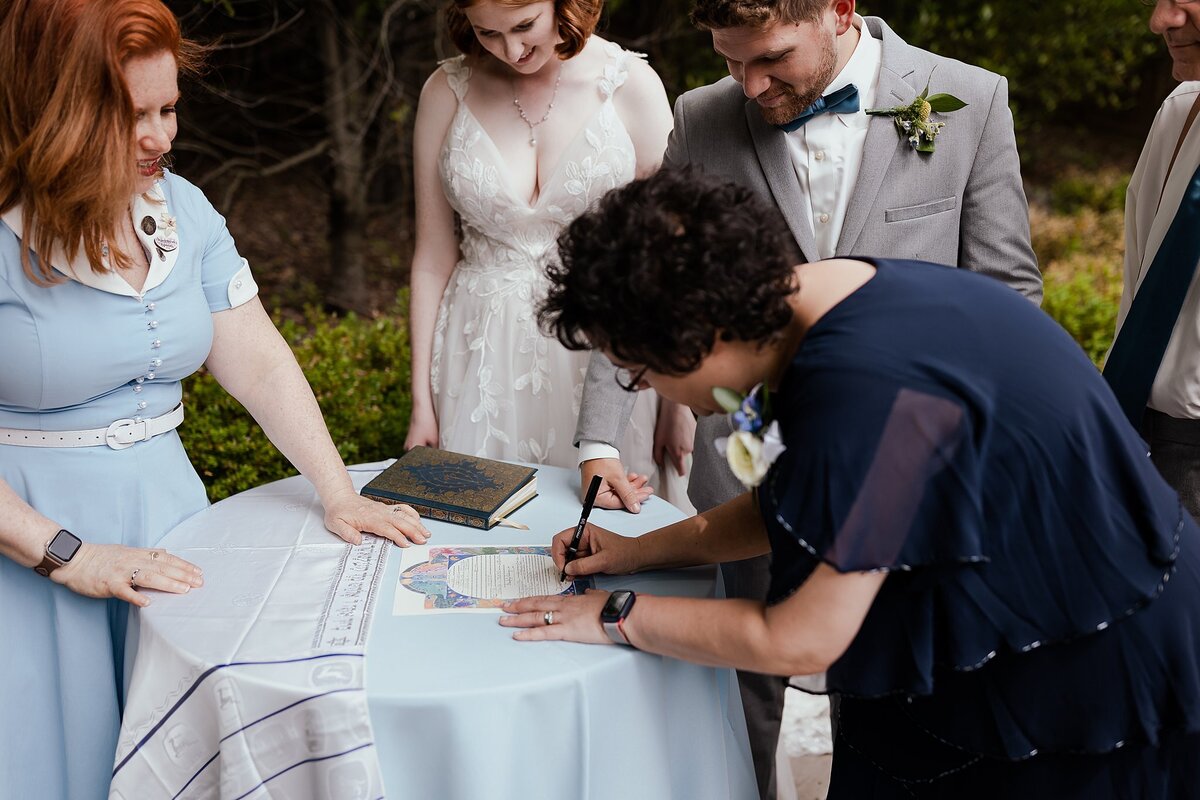 The red haired officiant stands beside the bride wearing a lace wedding dress with a plunging neckline and the groom wearing a light grey suit as they watch the mother of the groom signs the ketubah while wearing a navy blue dress. The groom's tallit is on the table beside the ketubah at this Jewish Wedding in Nashville, TN