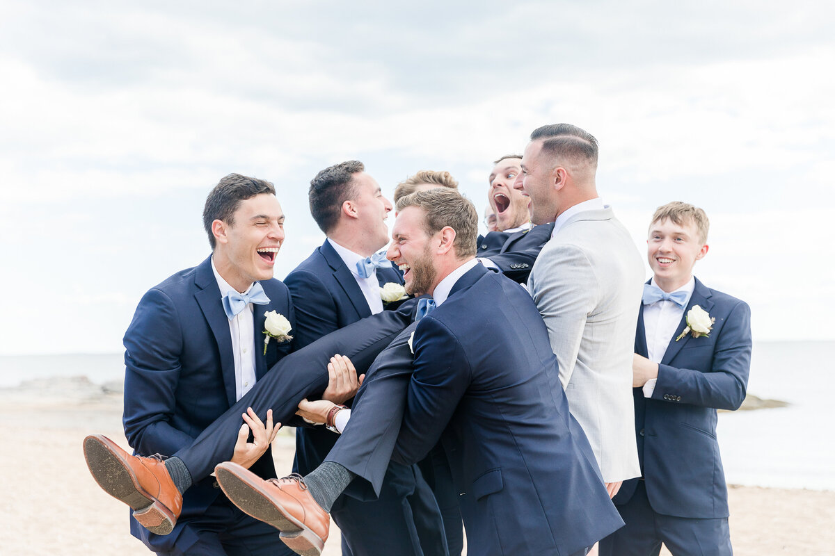 Groomsmen lift of up the groom on the shoreline of the Madison Beach Hotel. All of them are laughing. Captured by best New England wedding photographer Lia Rose Weddings.