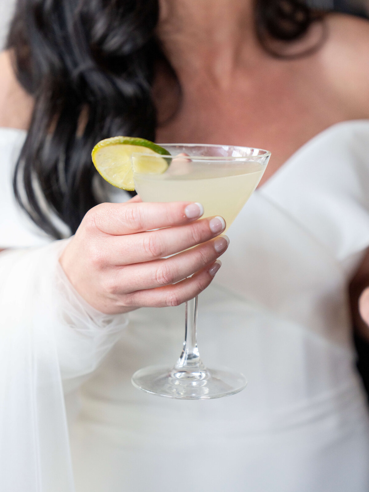 A person in a white dress holds a martini glass with a light-colored drink, garnished with a lime slice.