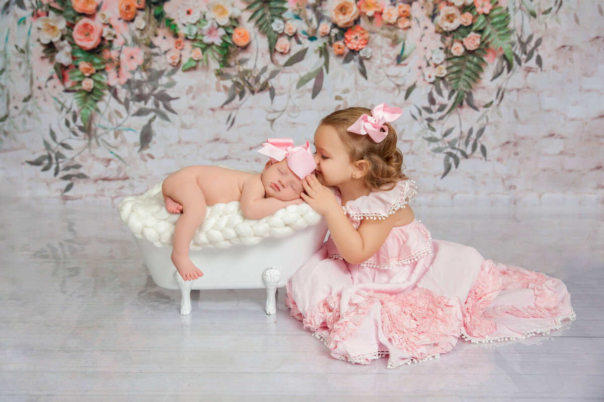 A young girl in a pink dress kisses a baby lying in a small white bassinet, both with matching pink bows, against a floral backdrop—capturing the sweetness of siblings by a Jacksonville newborn photographer.