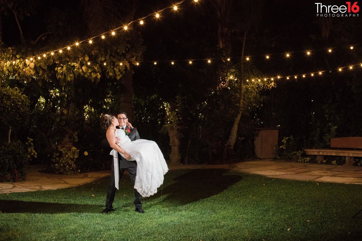 Groom carries his Bride under the lights of outdoor reception area