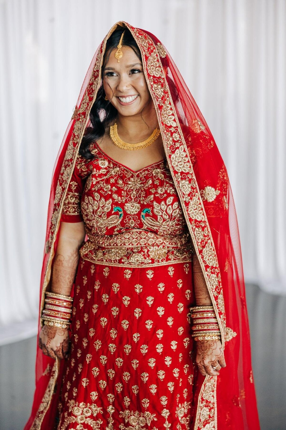 A smiling bride dressed in a traditional red and gold embroidered lehenga with her head covered by a veil.