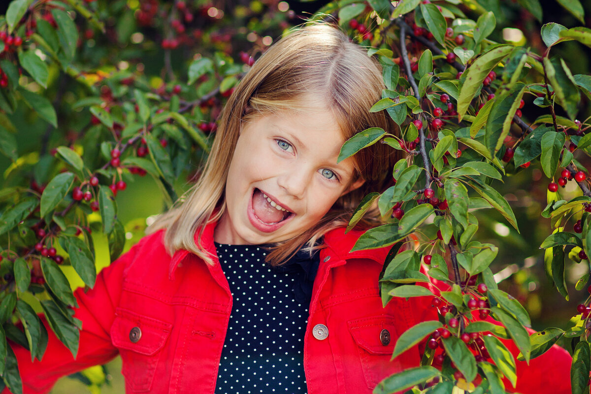 Young girl plays peek-a-boo through a bush that has red berries, matching her red jacket.