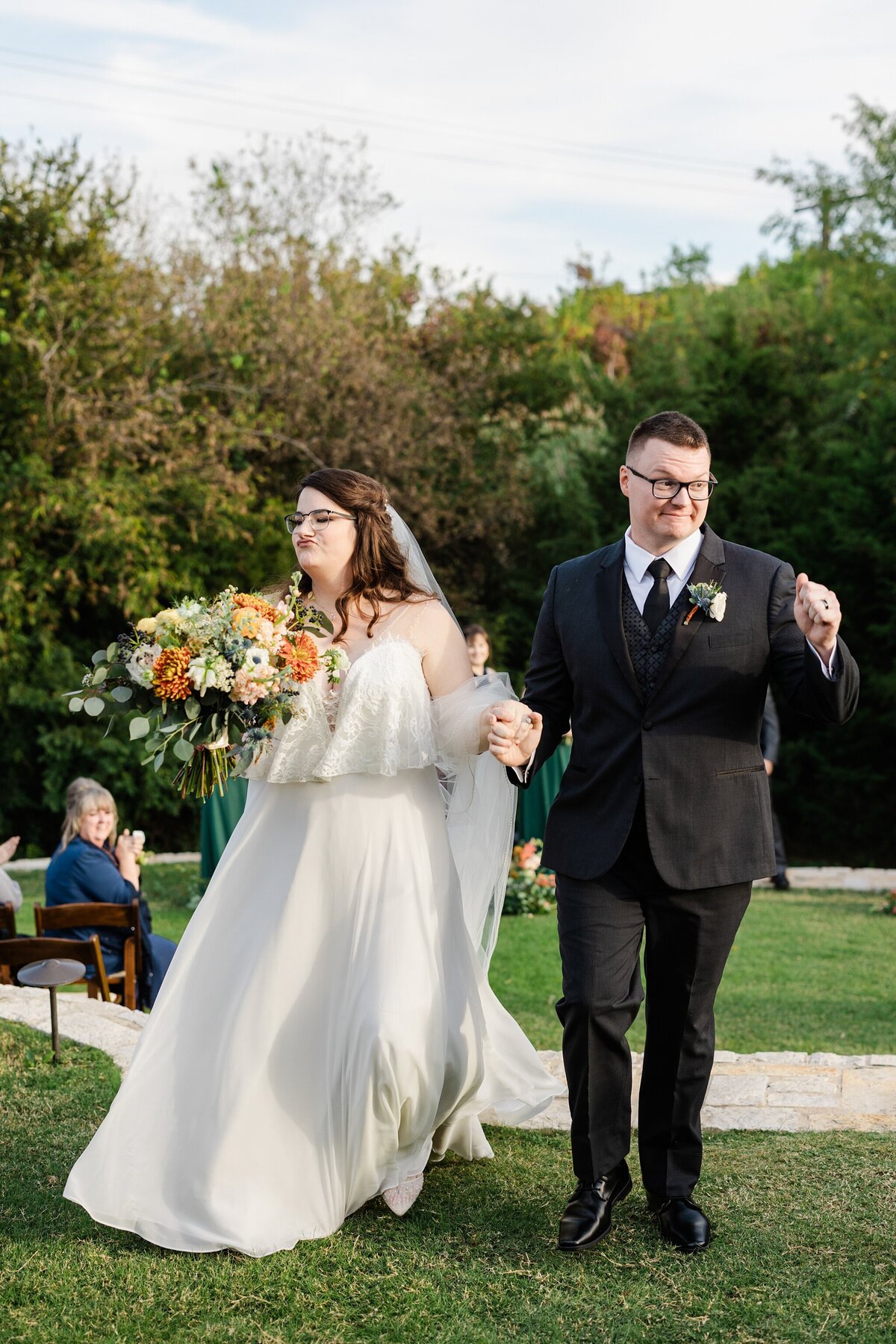 A candid shot of a bride and groom joyfully and jokingly recess down the aisle after their wedding ceremony at The Laurel in Grapevine, Texas. The bride is on the left and is wearing a long, flowing, white dress and a long veil while holding a large bouquet. The groom is on the right and is wearing a black suit with a boutonniere. They are both holding hands and wearing glasses.