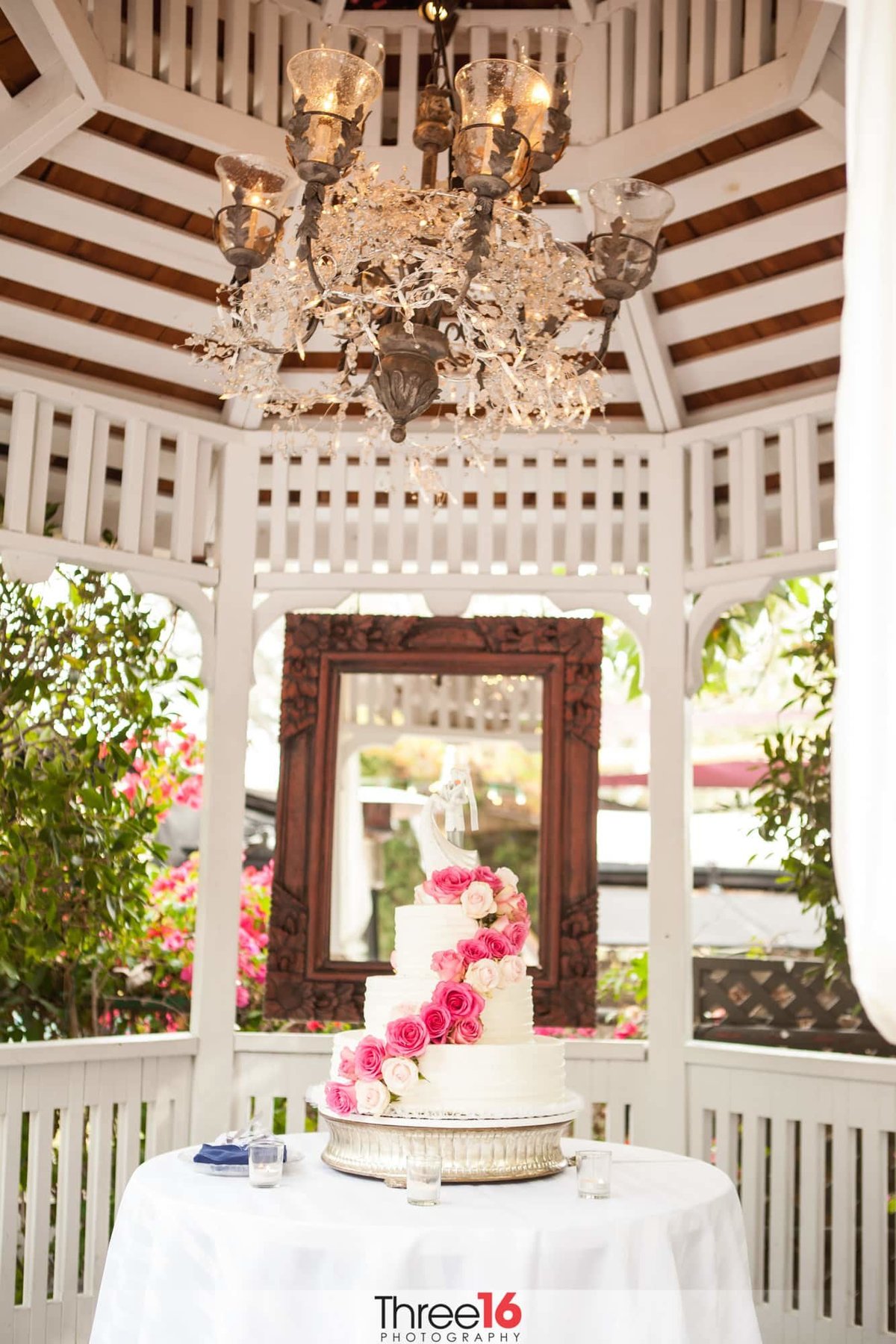 Beautiful 3-tiered white wedding cake with a string of pink flowers draped across it in a gazebo in front of a mirror