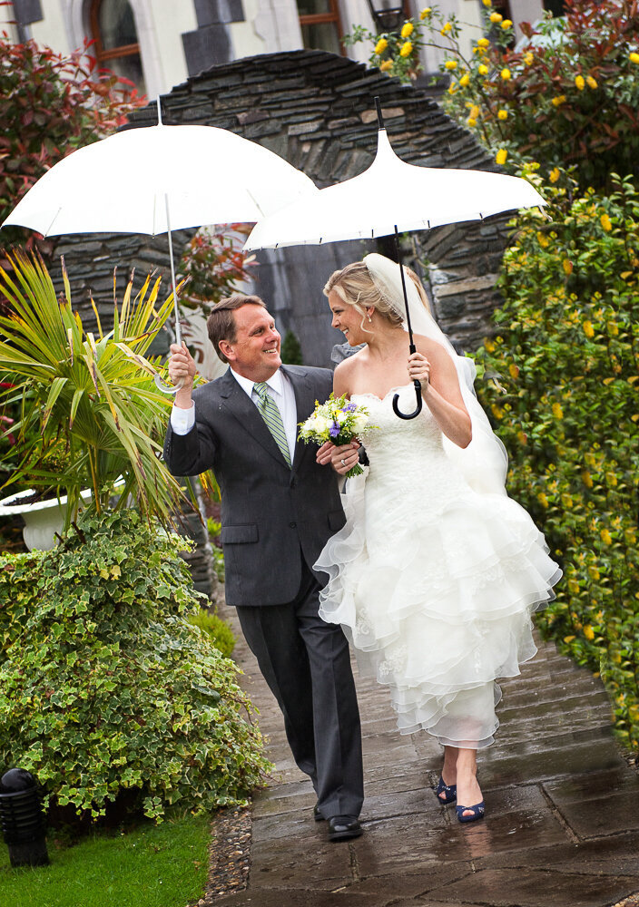 Bride wearing an A-line wedding dress with tulle ruffles and navy wedding shoes walking with her father, while holding white umbrellas in the rain at the Muckross Park Hotel