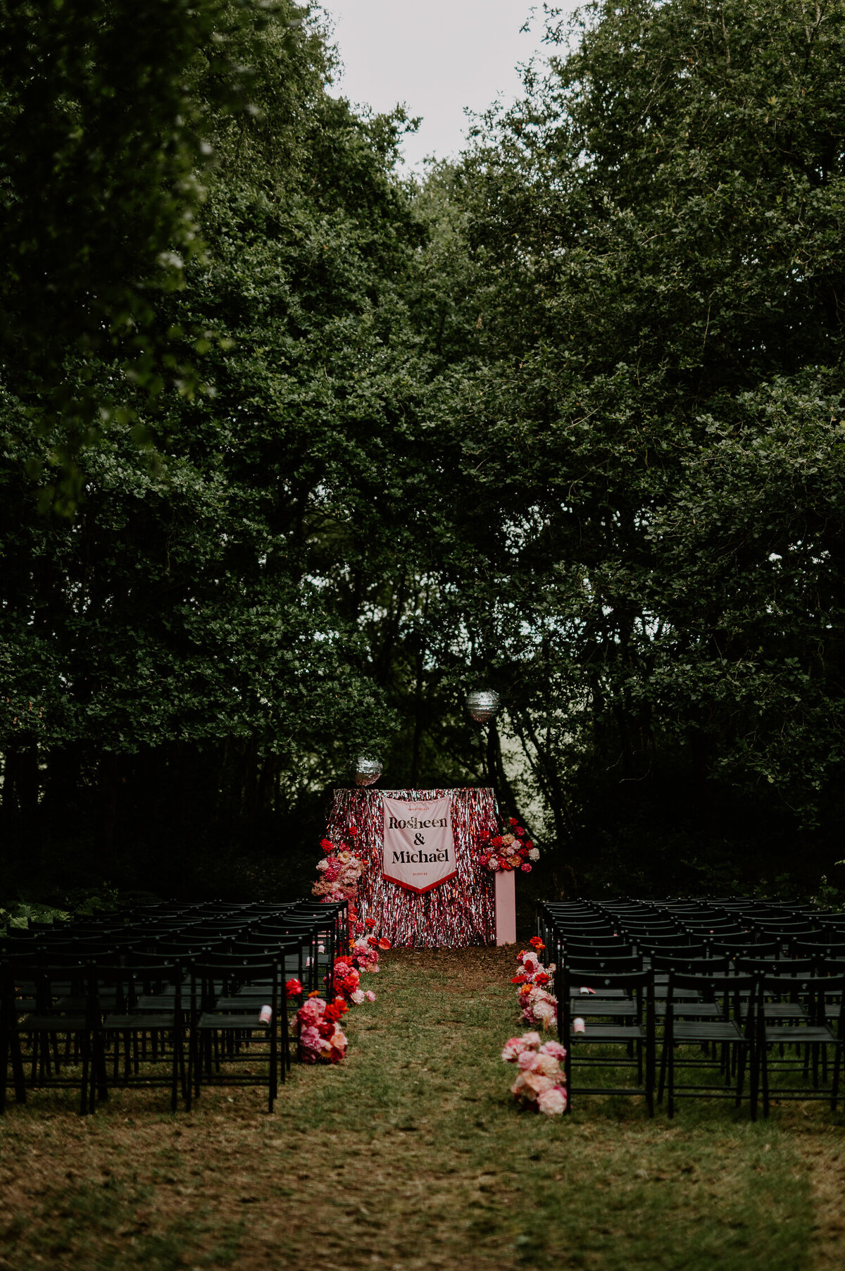 A pink woodland wedding in Coventry. Black chairs are lined up for guests with pink flowers at the end of each row. At the top of the aisle is a pink streamer backdrop with a pink custom wedding banner.