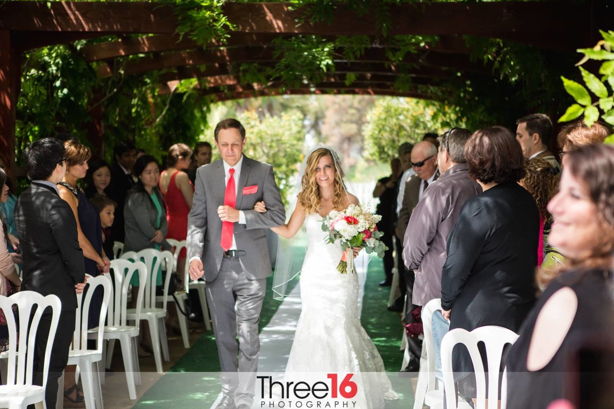 Bride is escorted under the foliage tunnel aisle leading to the altar and her awaiting Groom