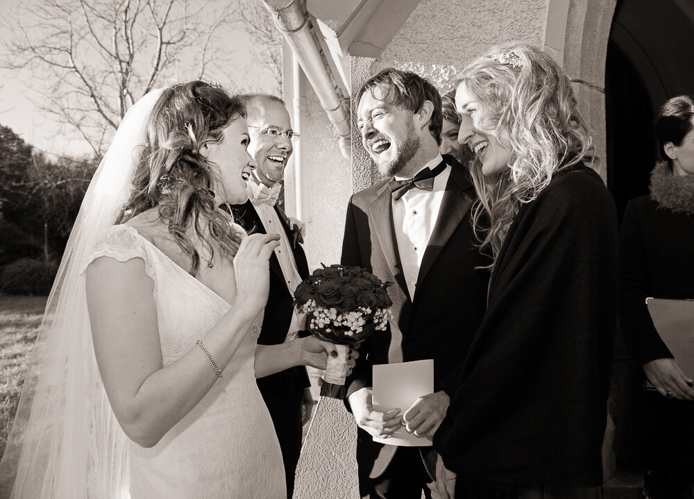 bride wearing a capped, fitted wedding dress with groom wearing a white tie, coattail suit laughing with friends outside Kenmare church