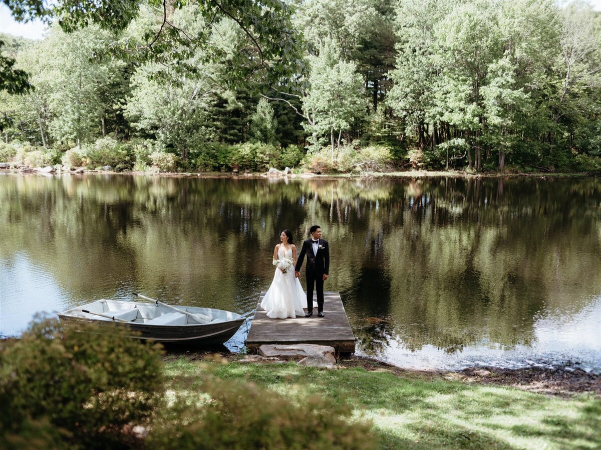 Stunning wide angle photo of bride and groom standing on wood dock next to old rowboat in front of calm lake and forest at Cedar Lakes Estate wedding venue