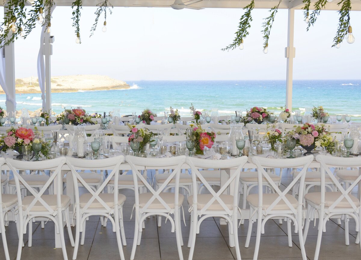 White chairs line a reception table decorated with fresh flowers  against a clear blue sea