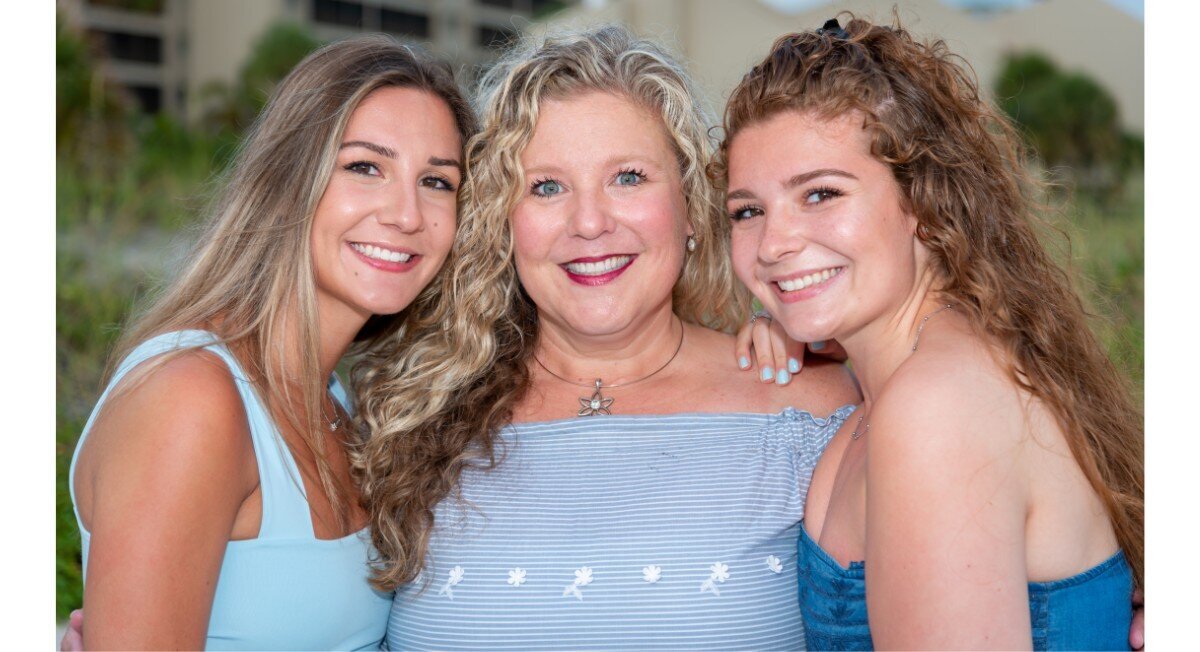 Three women with curly hair pose close together in front of a building on the beach for a family photograph.