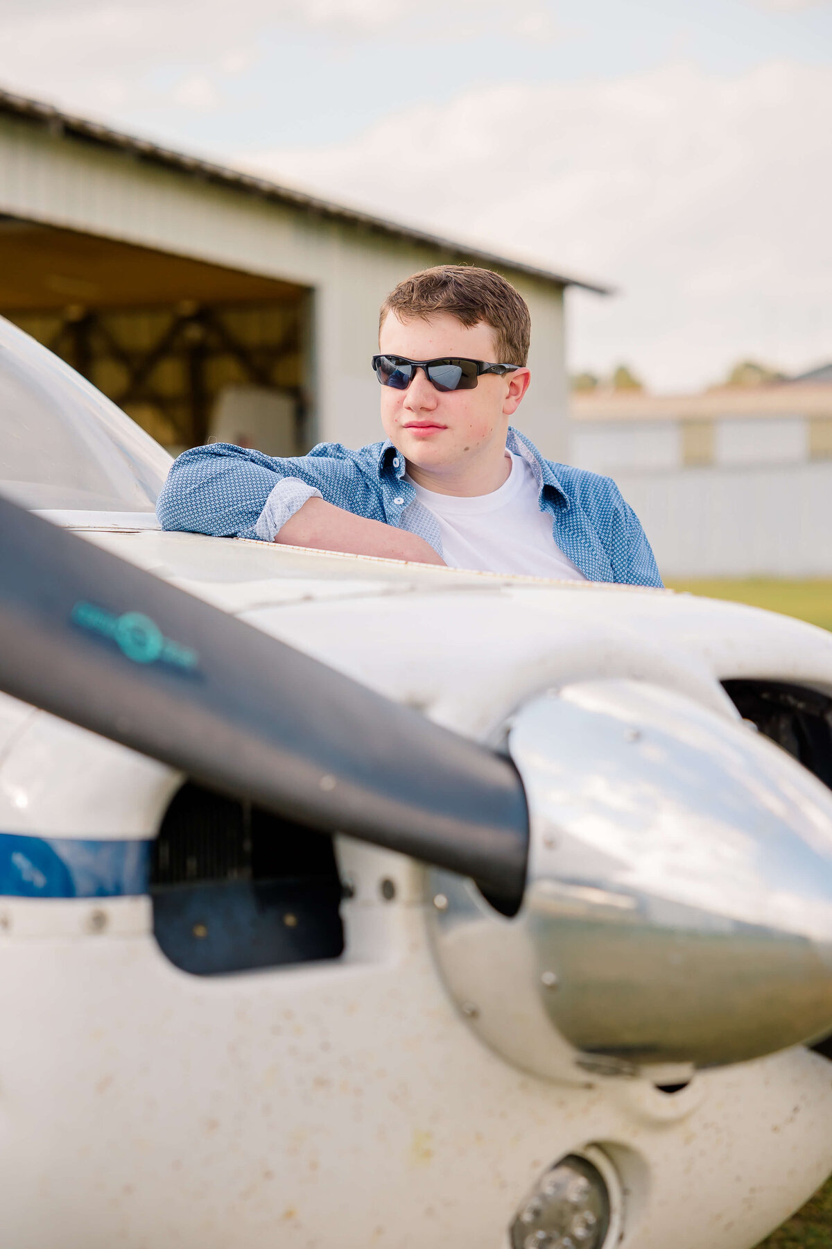 A senior photographer in Fayetteville, NC, wearing sunglasses and a blue shirt, leans against the nose of a small aircraft. The plane's propeller and engine casing are visible under a partly cloudy sky, with a hangar and grassy area in the background.