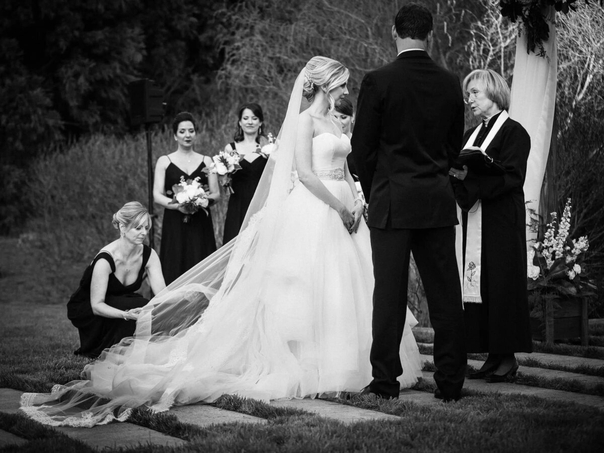 A black and white image of a bride and groom at the altar, with a bridesmaid adjusting the bride's veil, set against a serene outdoor setting.